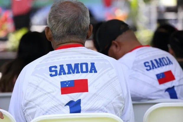 Two Samoan men, seated on folding chairs facing away from the camera, wear jerseys with the Samoan flag and the number one on the back.