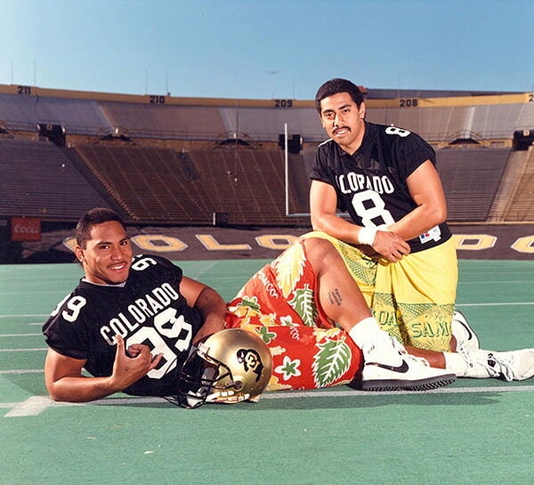 Okland Salava`e and Sal Aunese pose in football uniforms and lavalava on the field. Salava`e lies on the field. Aunese kneels next to him.