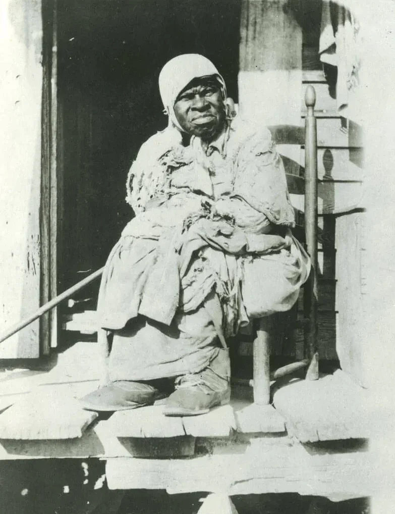 Grace Gates, an enslaved Black woman, wearing tattered clothing and a head covering, frowns as she sits on a chair on a dilapidated porch.