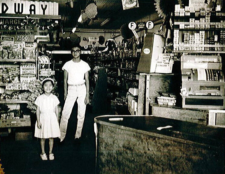 Chinese Americans Edward Woo, wearing a t-shirt and pants, and Nancy Gee, wearing a dress, stand inside his grocery store, Eddie’s Lucky Leadway.