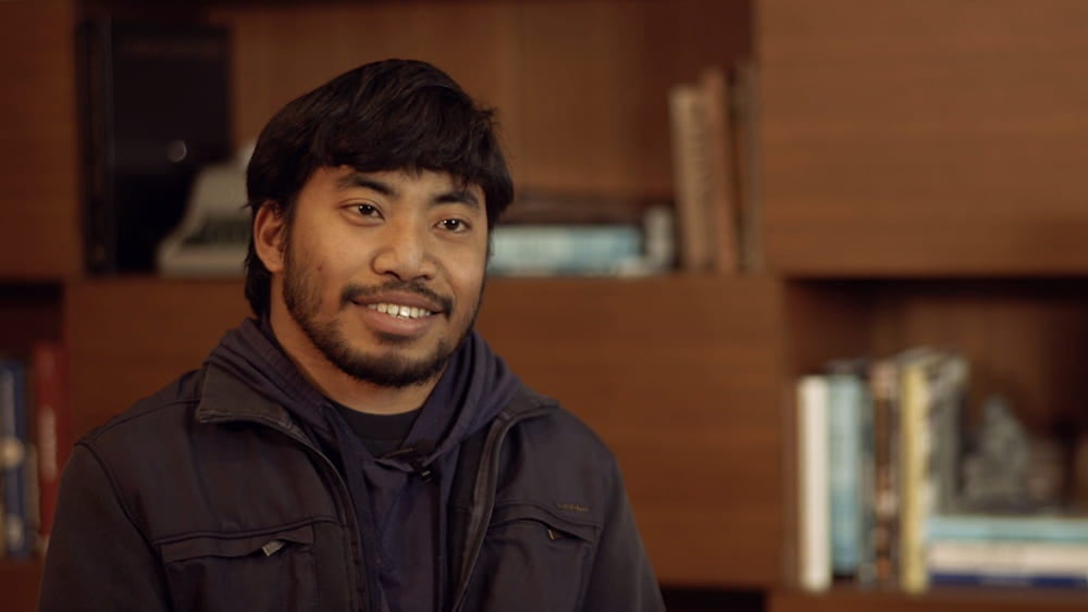 Headshot of Chris Balos, wearing a dark jacket and hoodie, who smiles while seated in office.