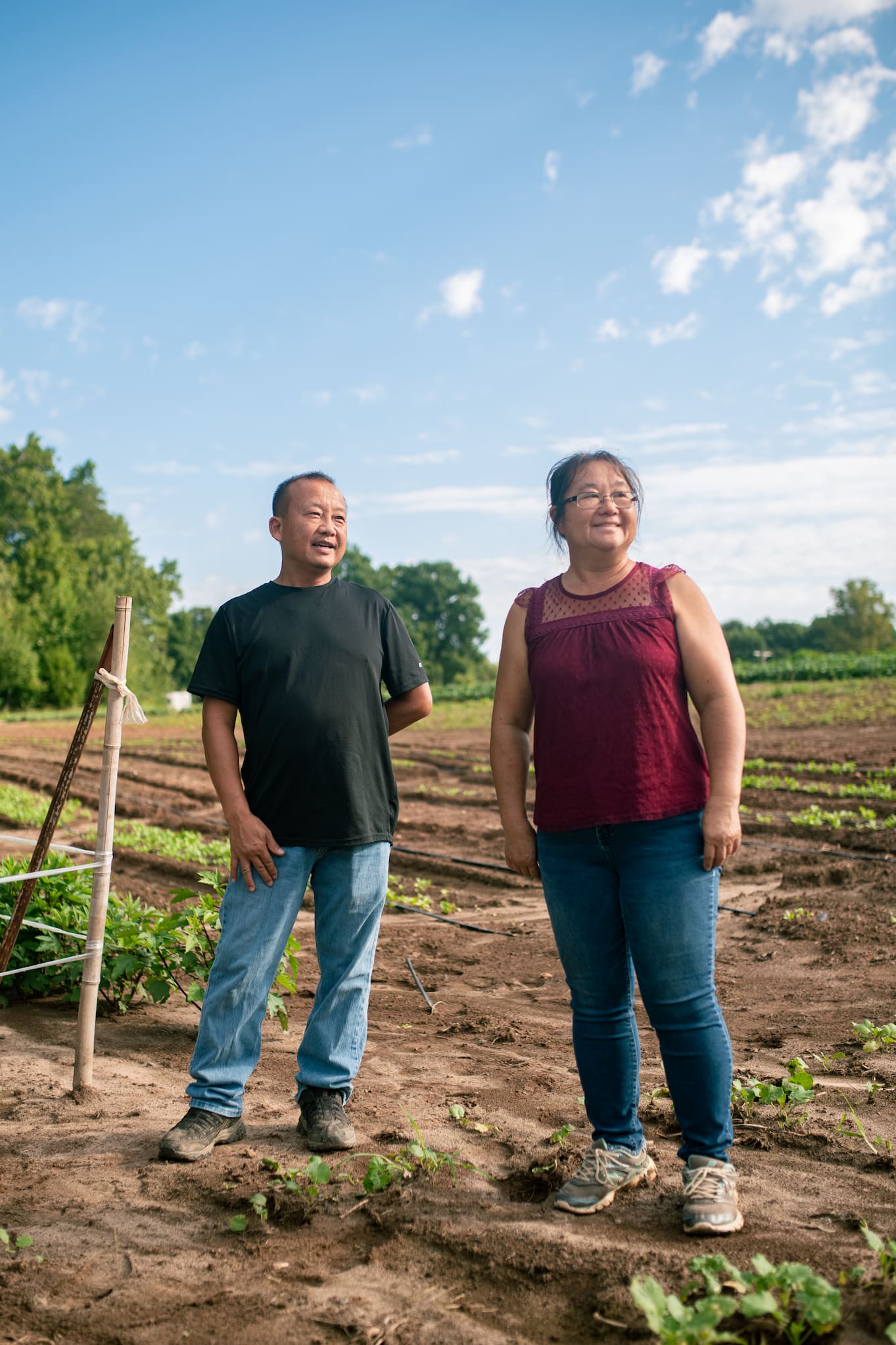 Hmong Americans Tou and Chue Lee of Lee's One Fortune Farm stand among rows of crops, casually dressed in jeans.