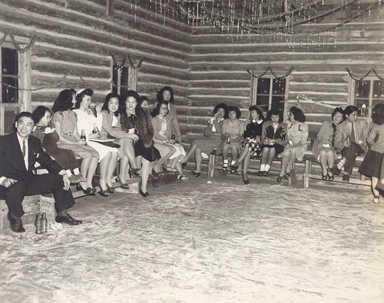 Chinese American young adults, wearing dresses and suits, sit together on chairs along the walls in a tinsel festooned segregated dance hall in 1946.