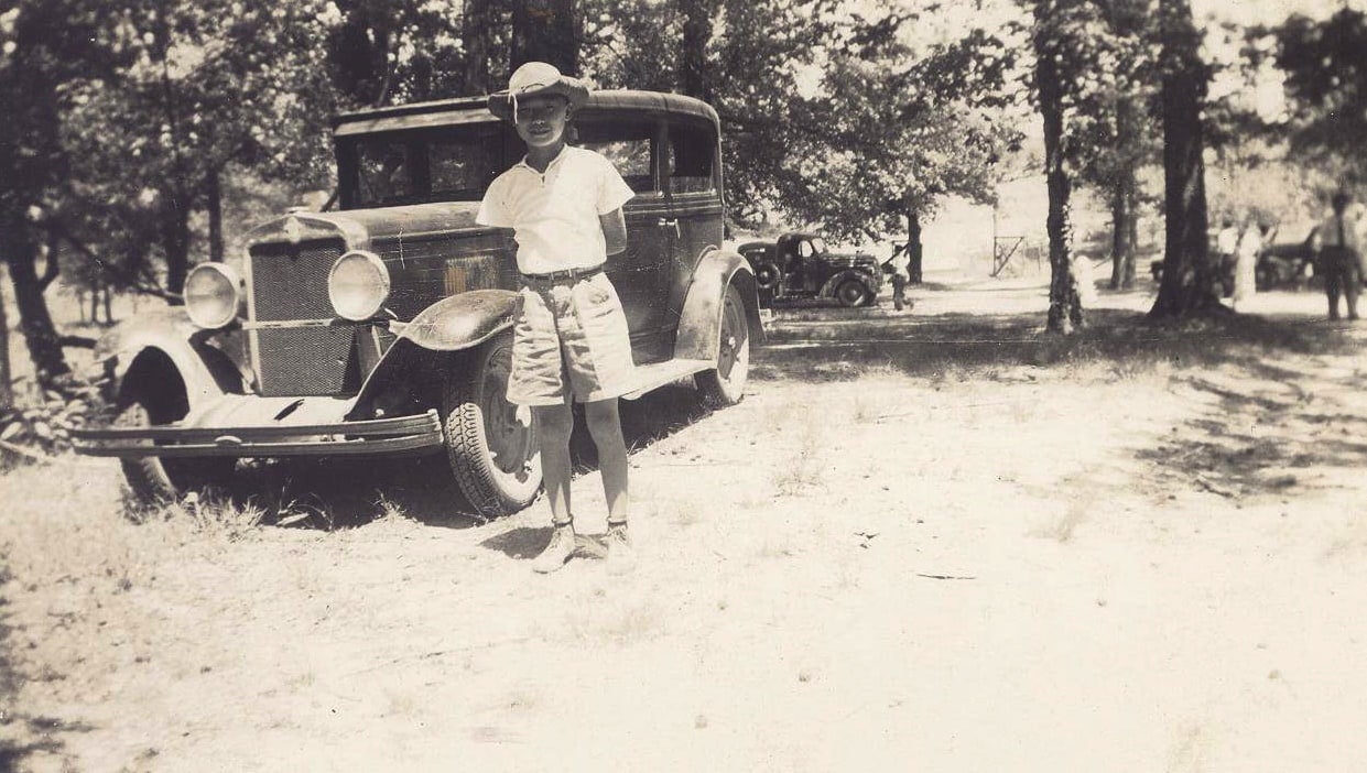 Chinese American Pershing Wong, wearing shorts, collared short-sleeved shirt and a hat stands in front of a parked car with his hands behind his back.