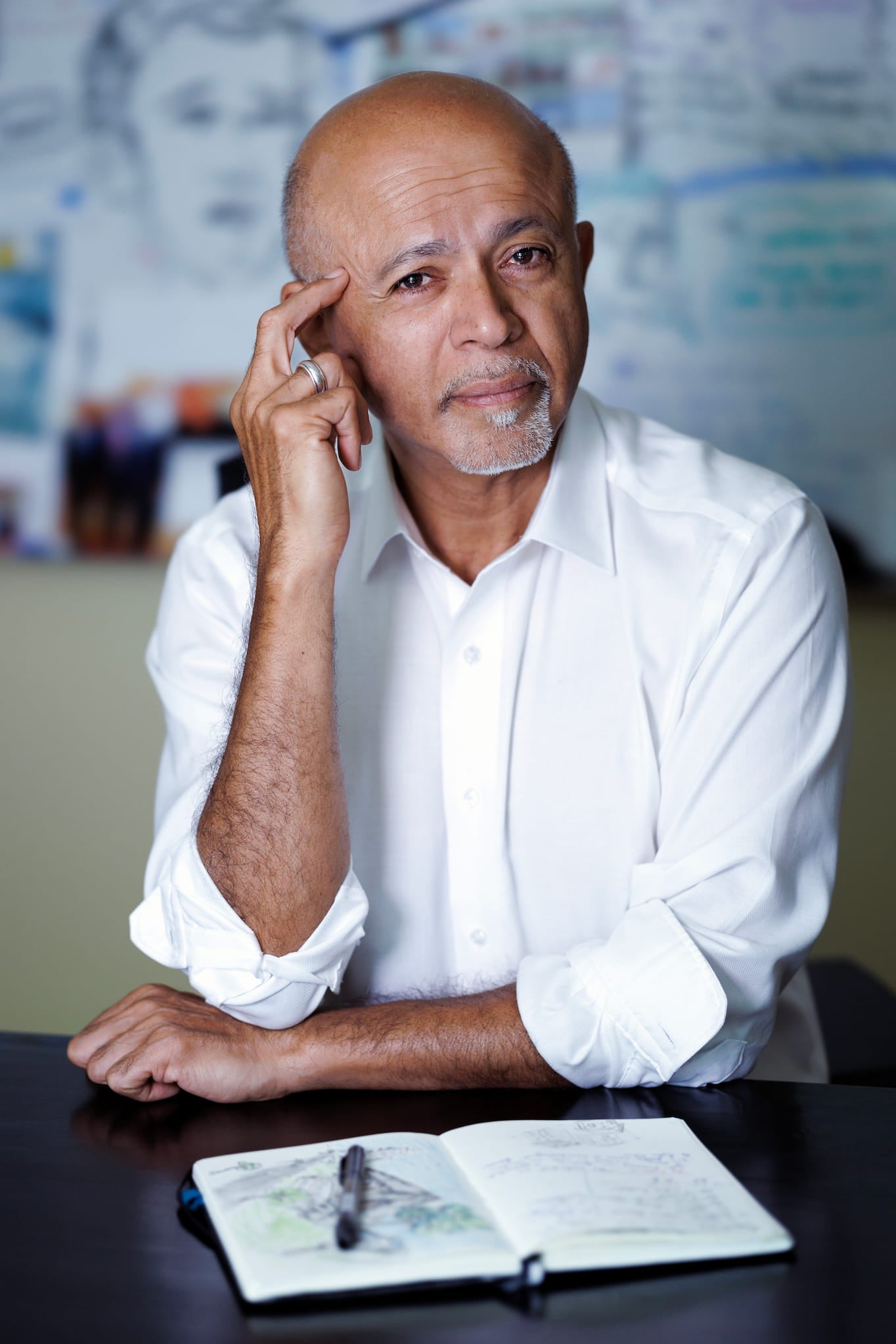 Dr. Abraham Verghese poses for a headshot wearing a button down shirt with a whiteboard behind him and an open notebook on the desk before him.