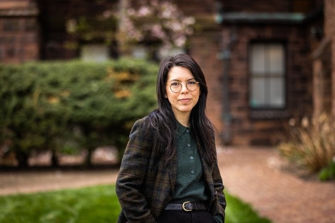 T Kira Māhealani Madden, poses for a headshot outside a brick building, wearing glasses, a tweed jacket and dark pants.