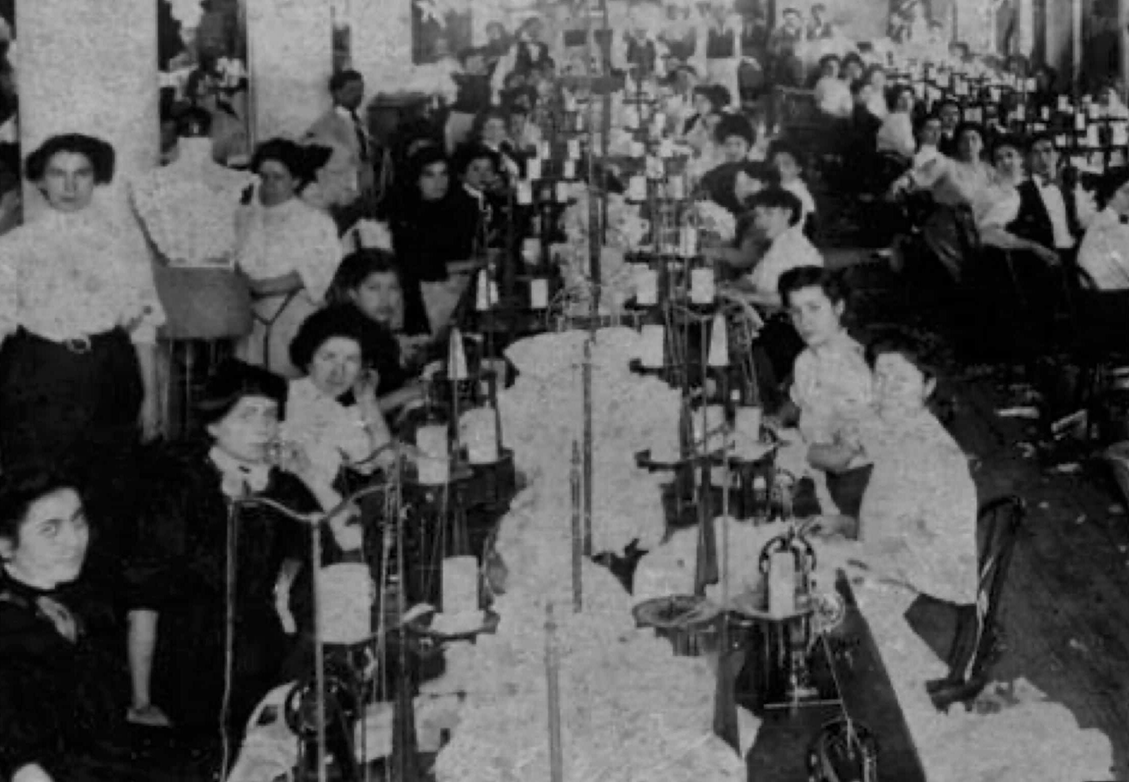 Rows of women sit at work stations in crowded garment factory.