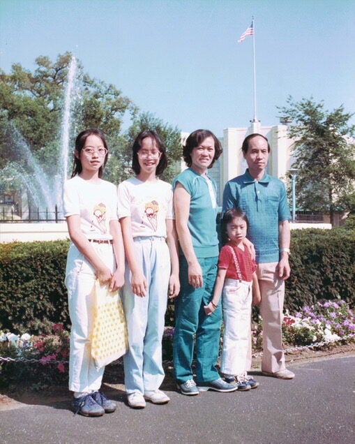 Mrs. Ma, a Chinese garment factory worker, poses with her family in front of a water feature and building with American flag.