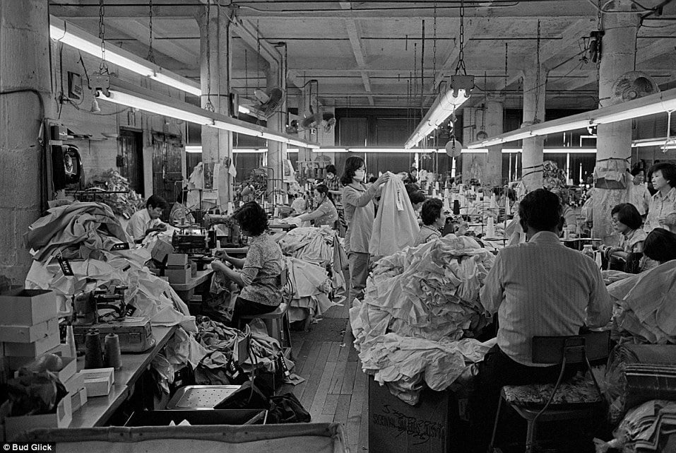 Interior view of crowded garment factory floor with workers surrounded by piles of clothes.