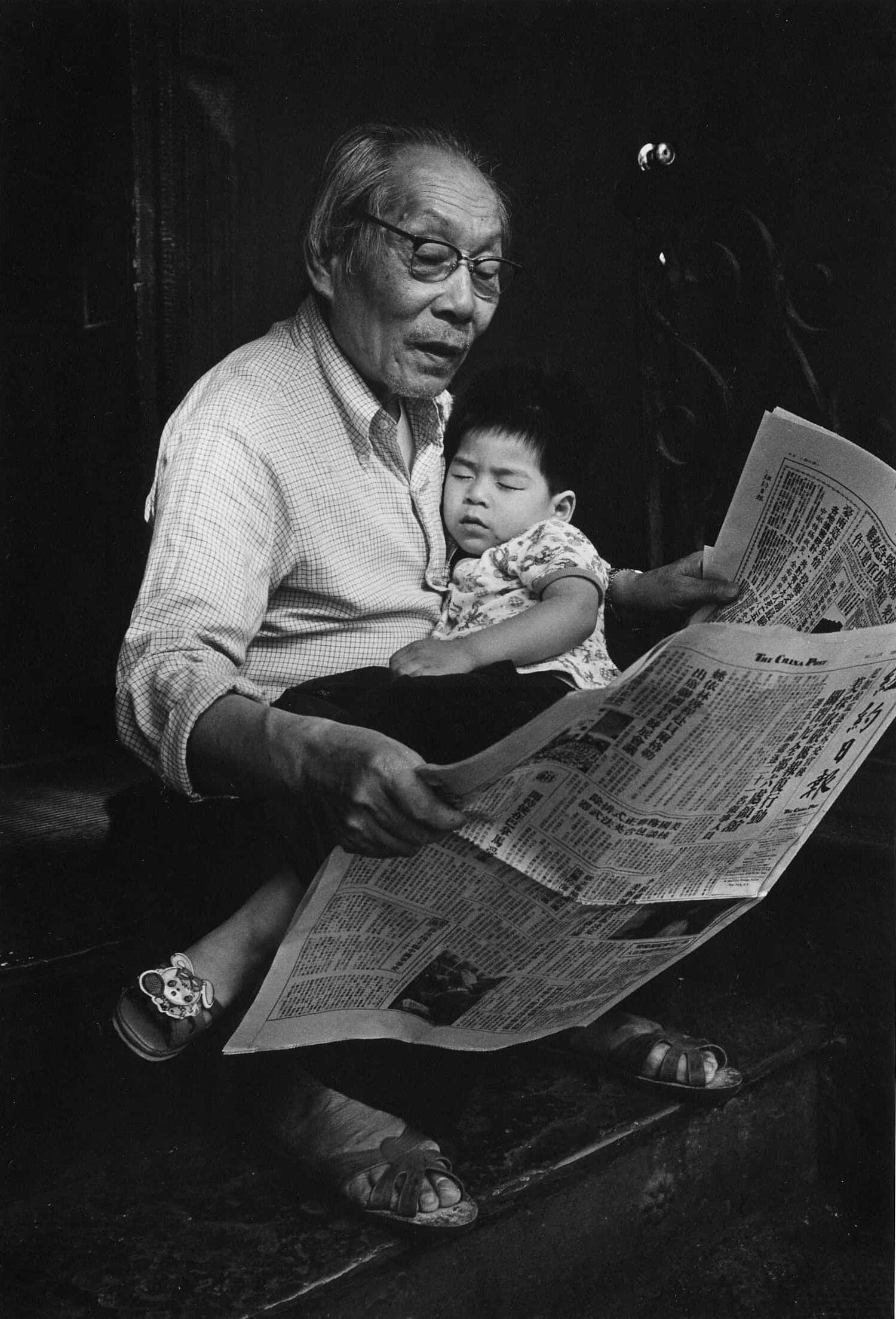 Older man holds sleeping child, his grandson, in his lap while reading a newspaper.