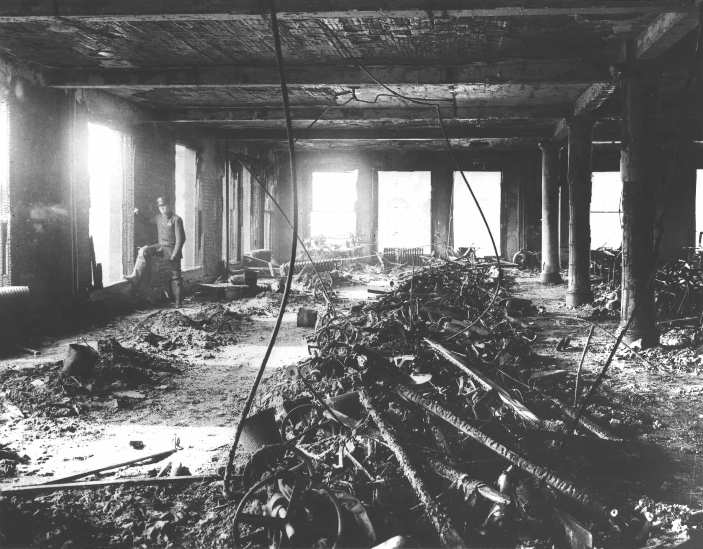 Burnt rubble and debris at the center of a factory floor shows the aftermath of Triangle Shirtwaist Factory fire. Man in uniform stands on perimeter of floor, near open window.