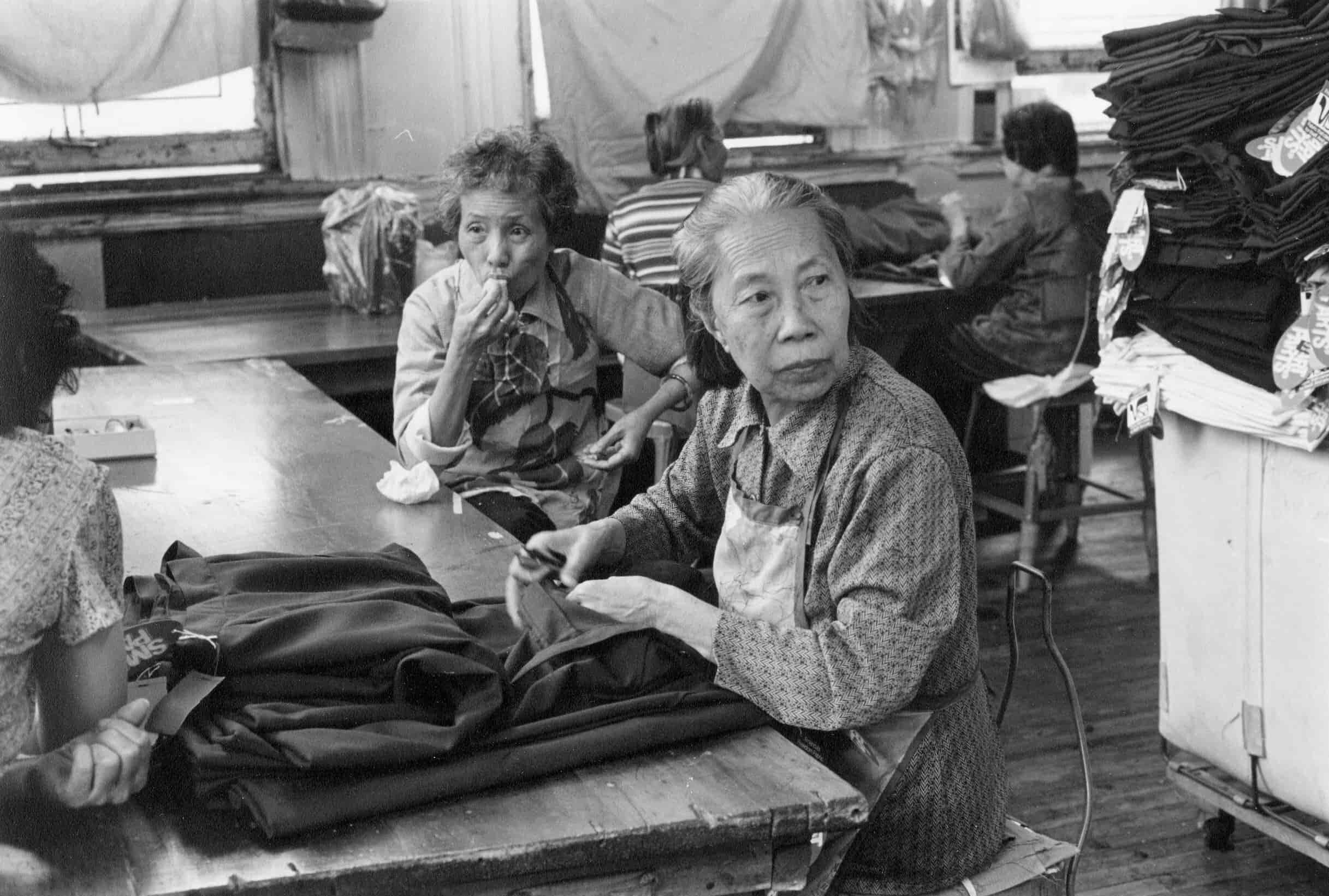 Elderly Chinese women sit at factory table. One worker has fabric in front of her, while the other eats a snack.