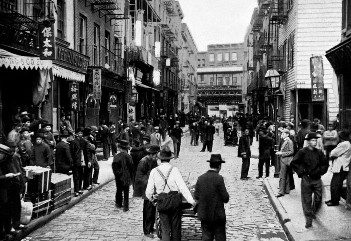 View of street in Manhattan's Chinatown. The sidewalks are crowded with men on either side of the street, with signs above written in Chinese.