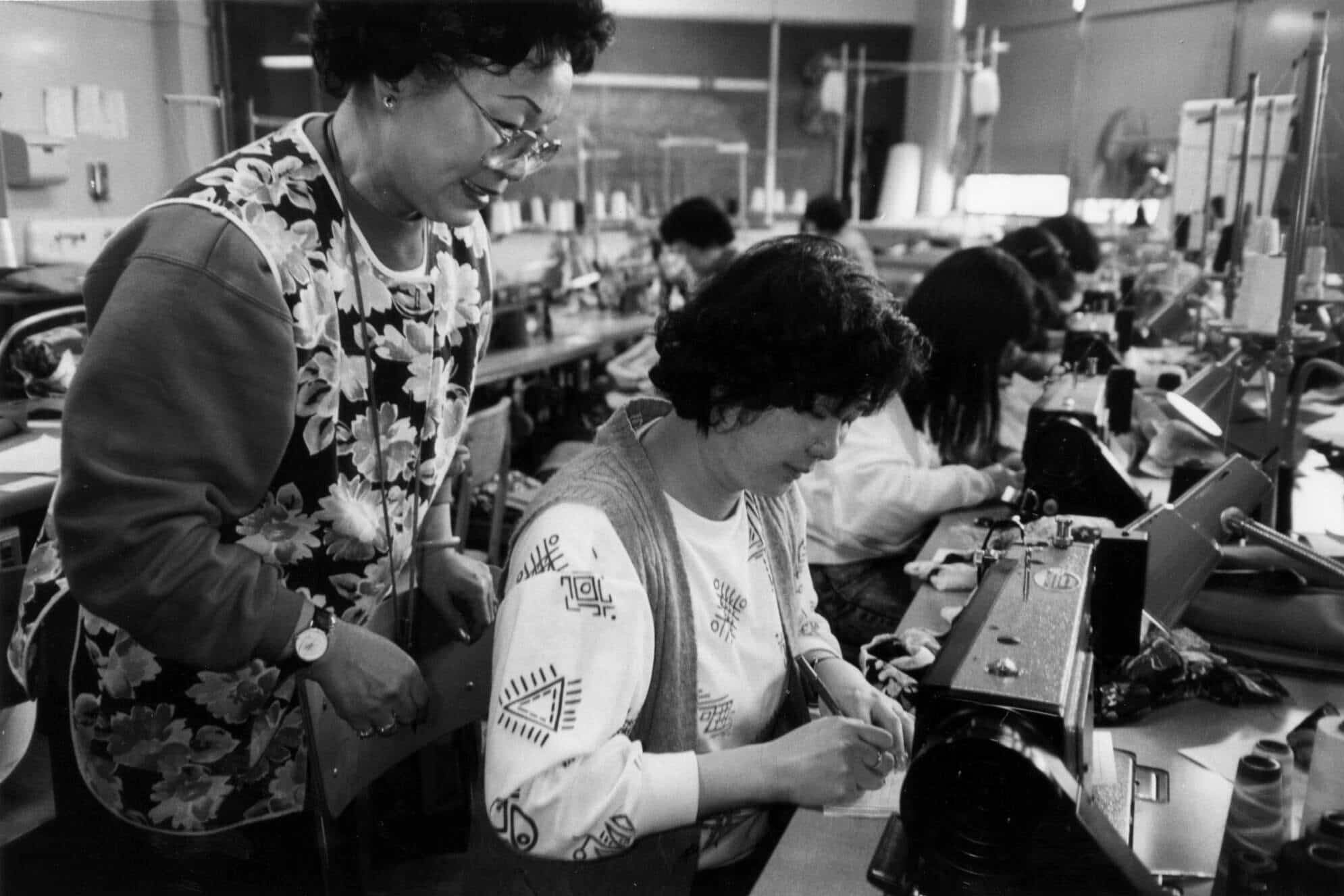 Factory worker Yook Chee Hom, seated in front of sewing machine, demonstrates a new sewing style to her factory co-worker, who stands beside her.