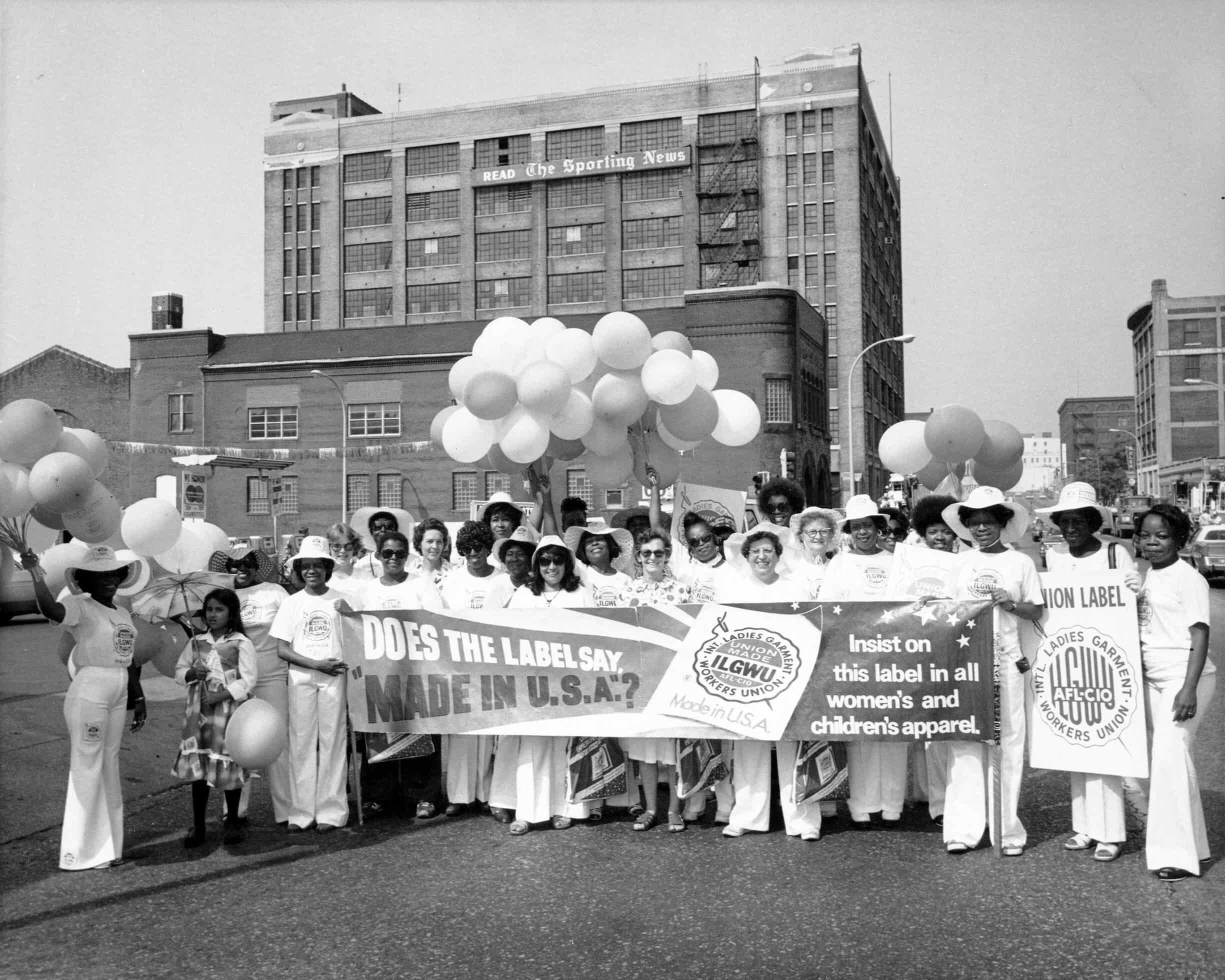 Group of ILGWU members stand outside of building. They hold a banner reading, "Does the label say "Made in the U.S.A."?"