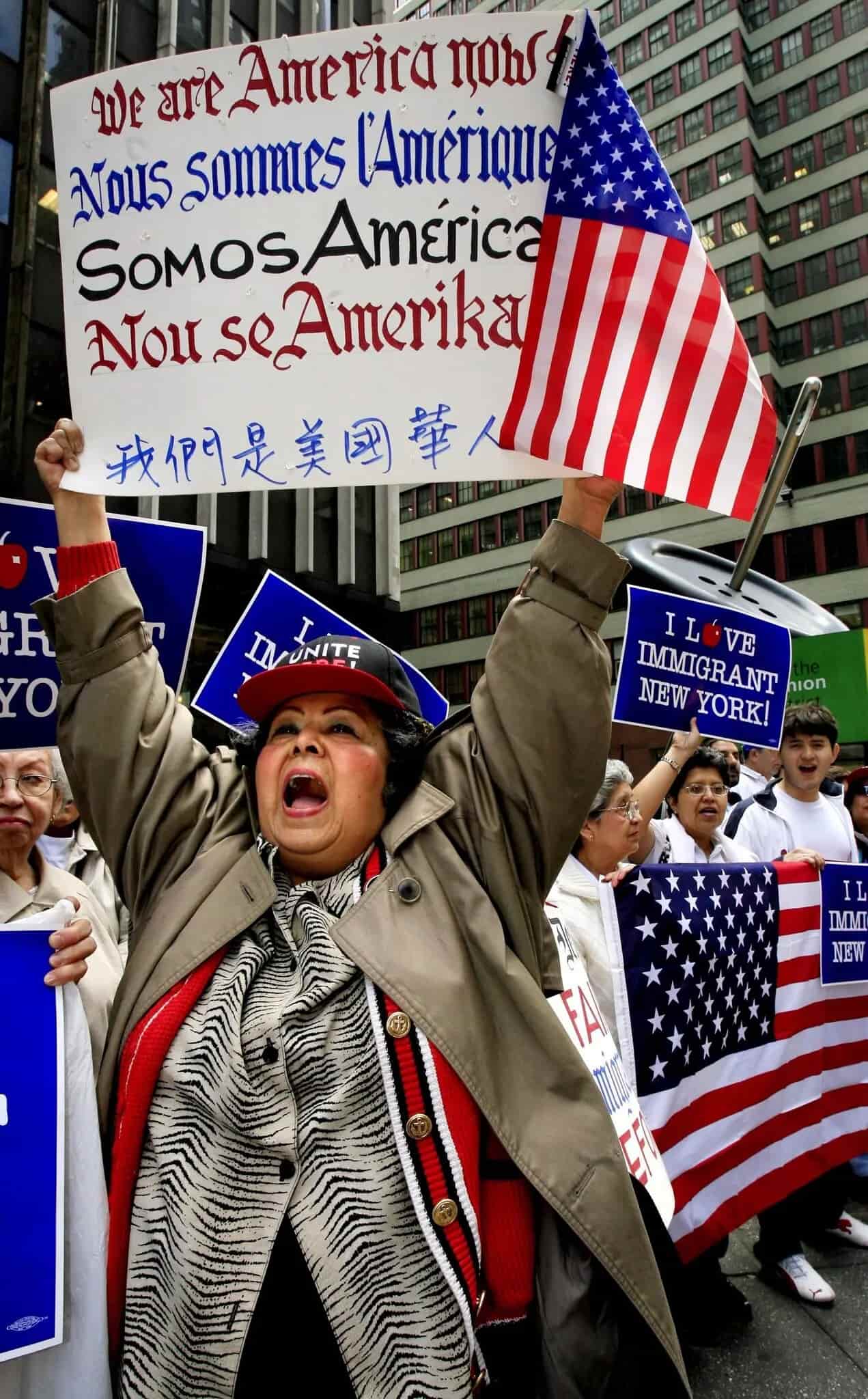 Kathy Andrade, woman in brown jacket, holds up a sign at outdoor rally for immigrants' rights. Others stand with "I Love Immigrant New York" signs.