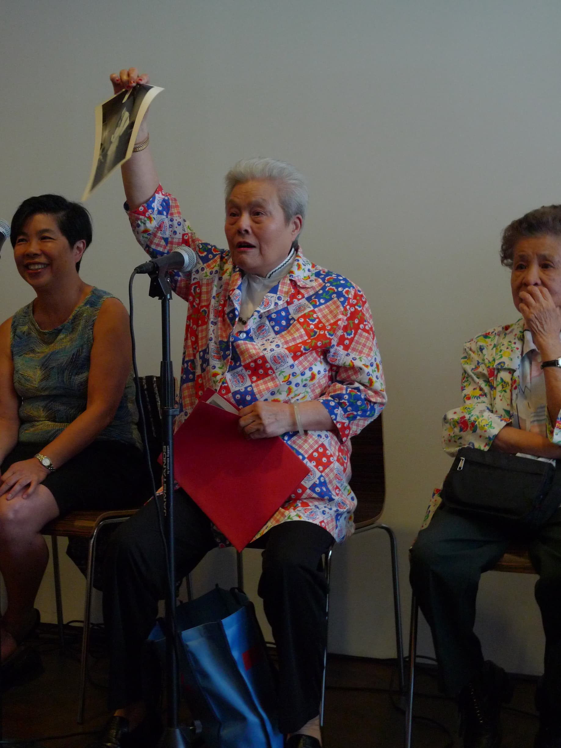 An elderly woman in printed jacket sits in front of a microphone with arm raised. Two women sit on either side of her.
