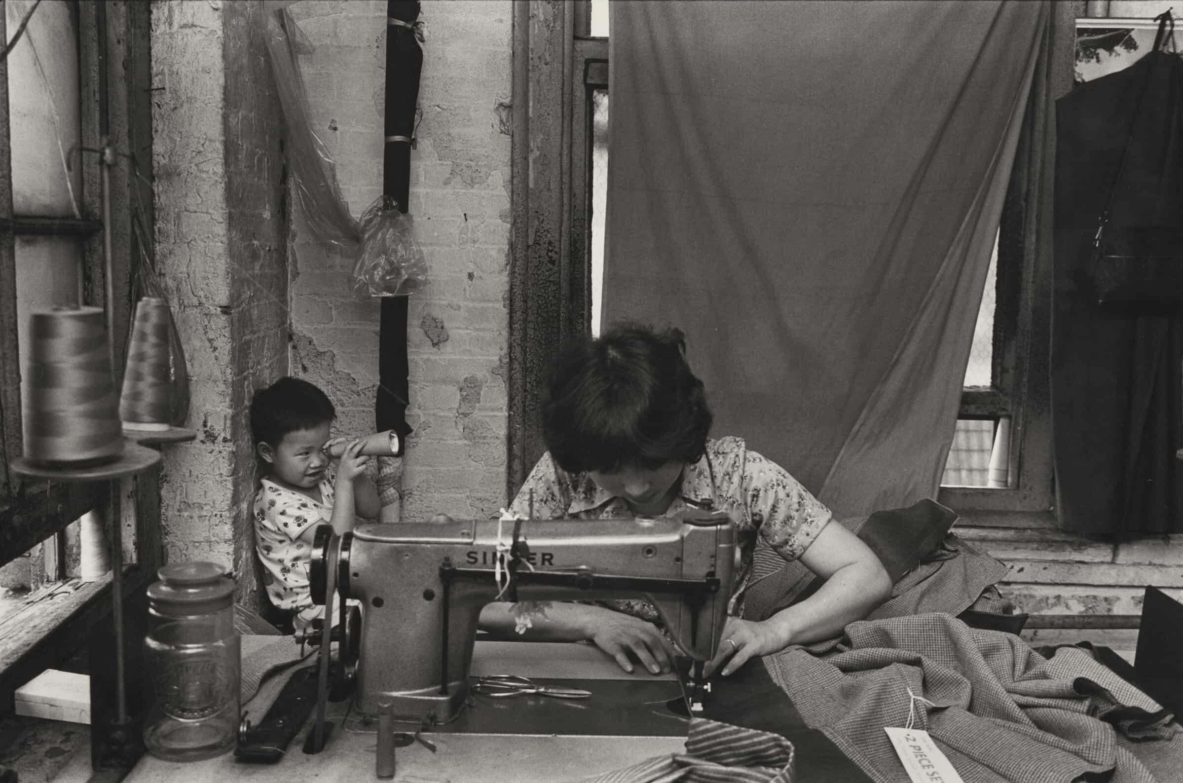 A factory worker at sewing machine. Her child, a young boy, plays in corner behind her.