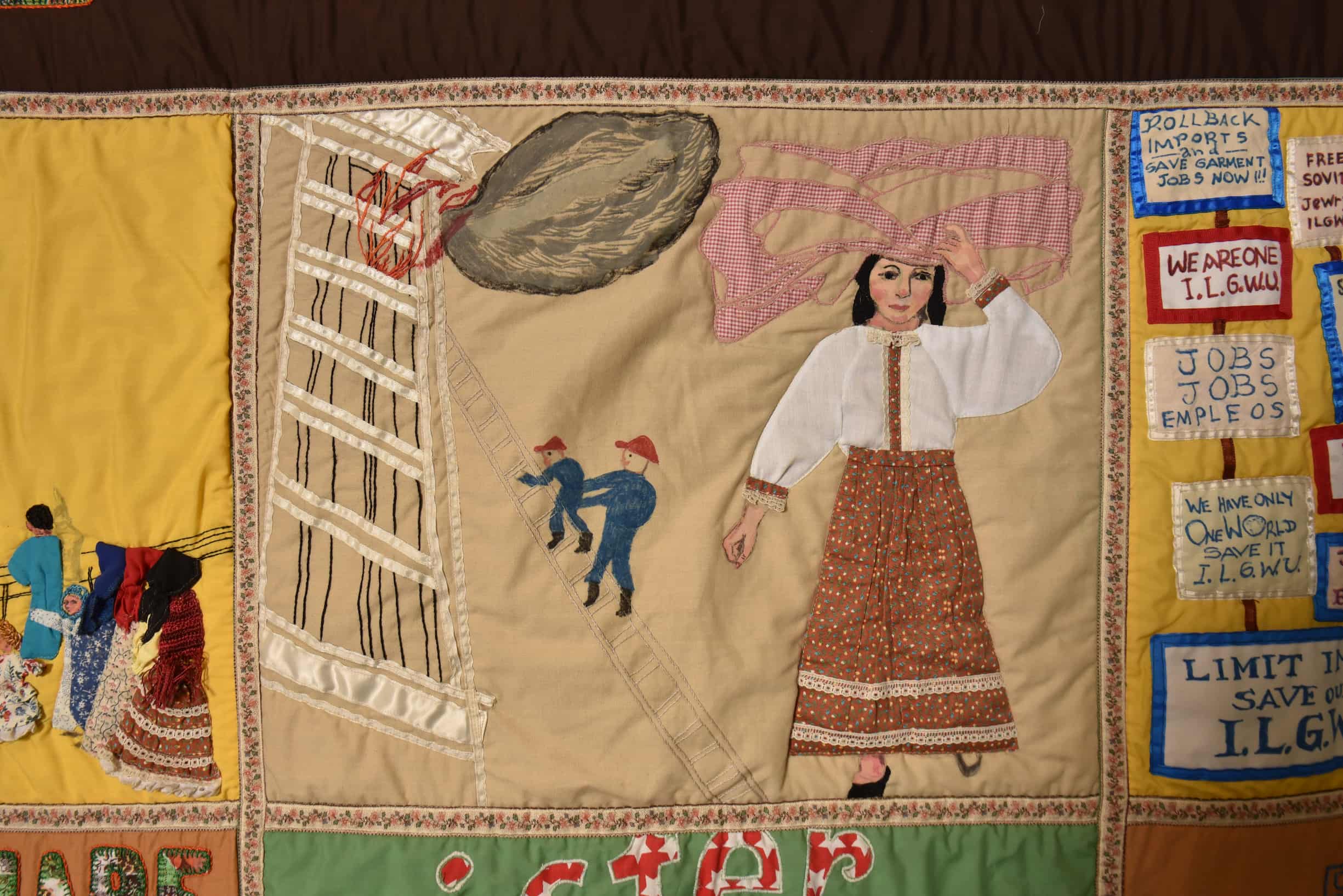 Close up on quilt square with tan background. A woman stands to the right, while firefighters climb up ladder into burning building on left.