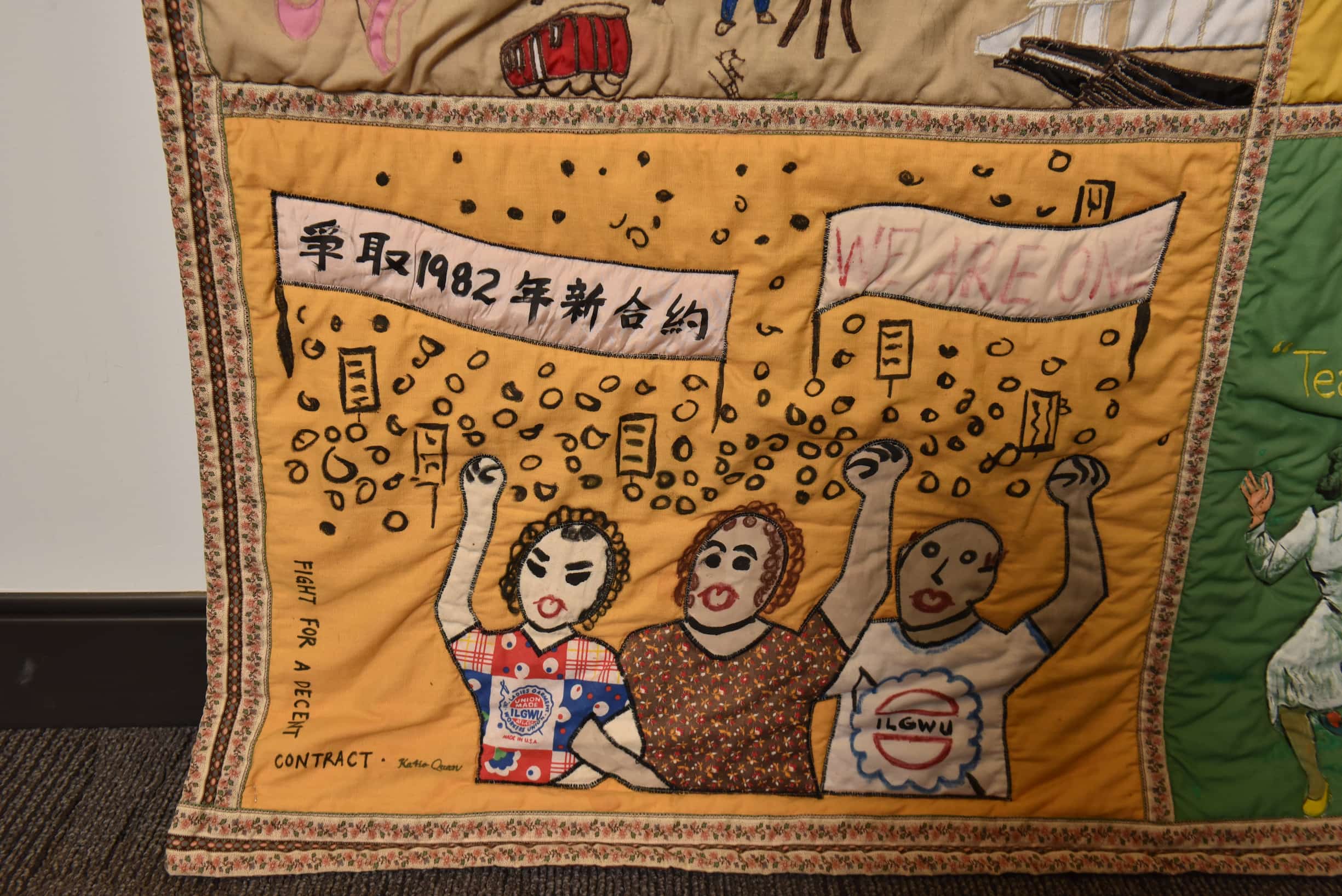 Close up on quilt square with orange background. Trio of workers with raised fist and banners behind them represent 1982 strike.
