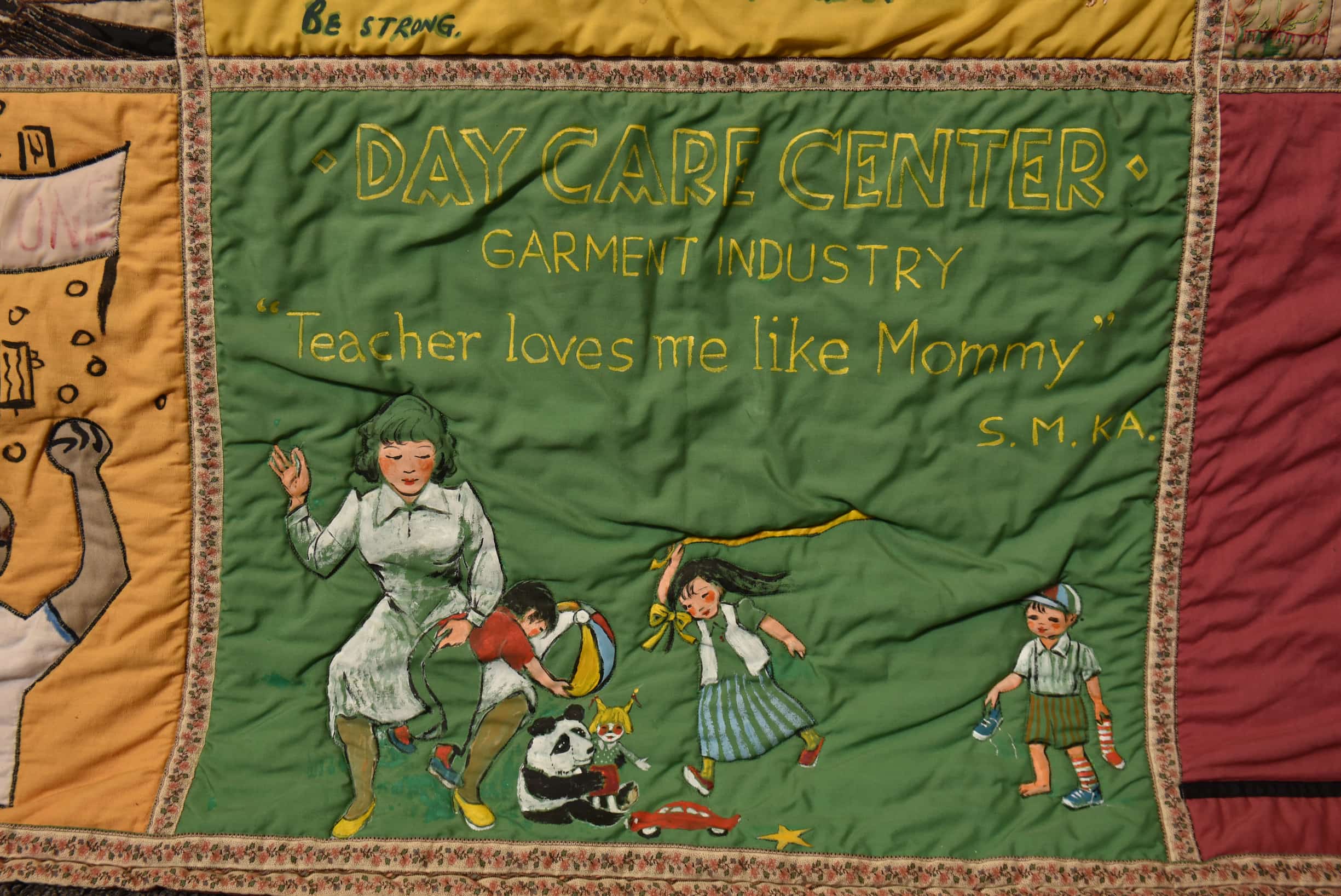 Close up on quilt square with green background. Text reads "Daycare Center" with female teacher disciplining child while two others play.