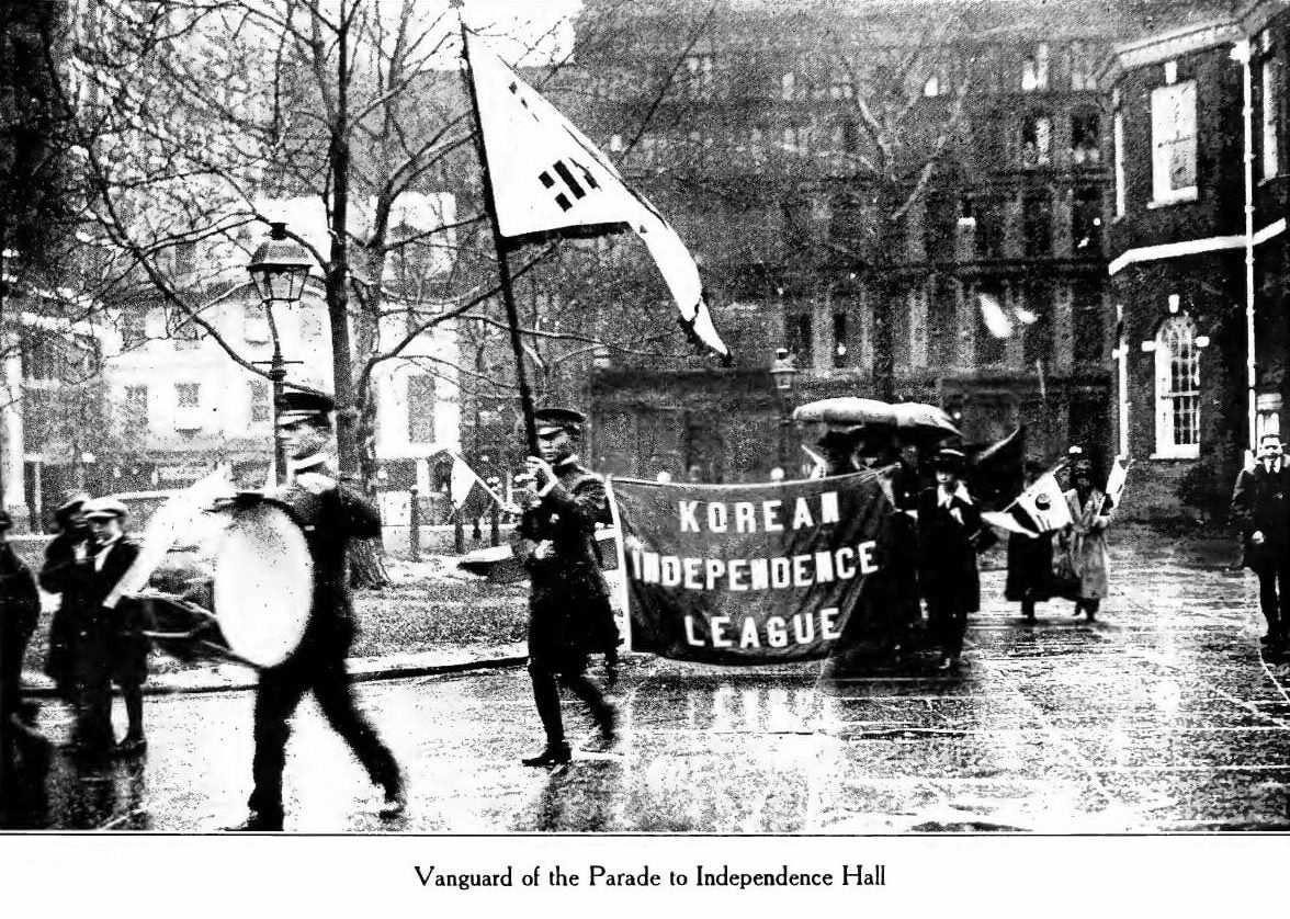 Small parade procession, with people carrying "Korean Independence League" banner, walk down street.