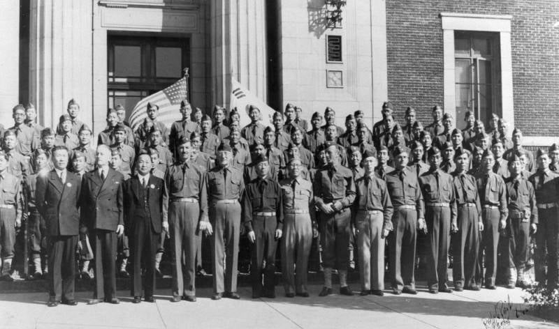 Uniformed Korean American men stand in rows outside of building. Flags of the U.S. and South Korea are seen behind last row of men.