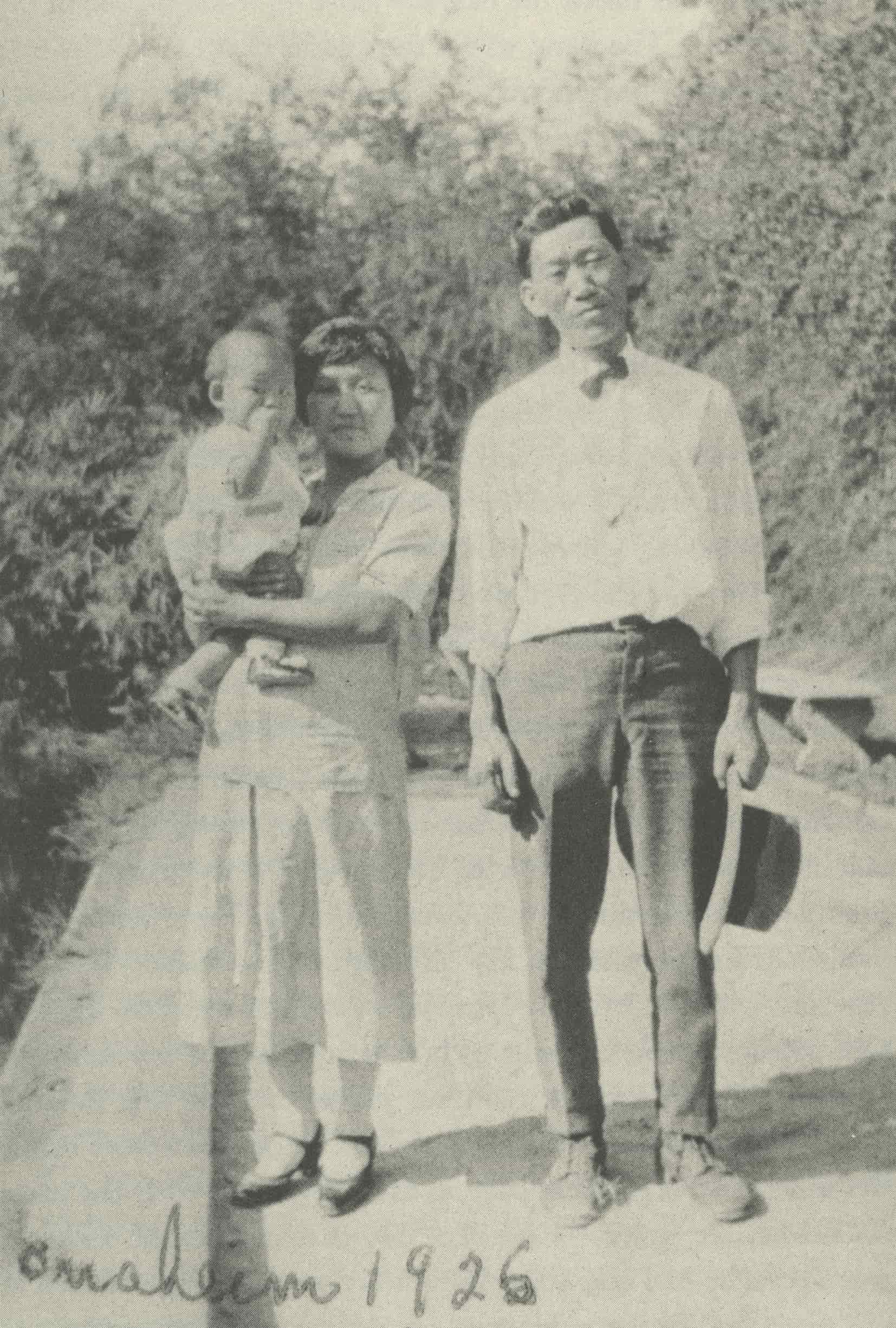 Korean American woman, Mary Paik Lee, carries her young son and stands next to her husband in a field.