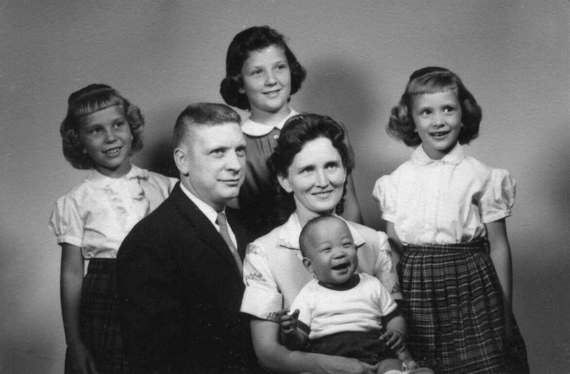White American couple are seated in portrait studio with their children. Their three daughters stand behind them, while the mother holds young son, an adopted Korean boy.