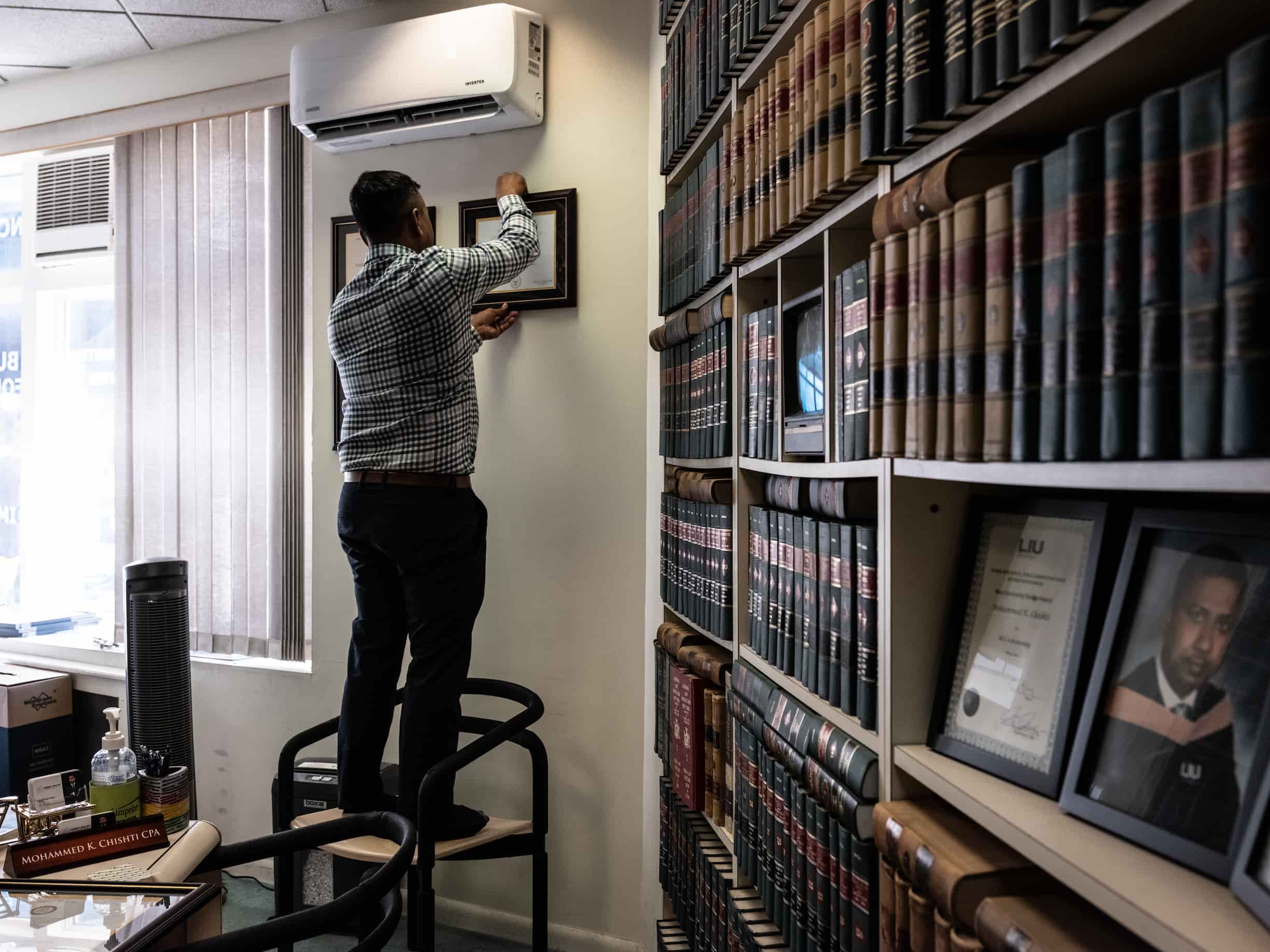 Bangladeshi American Mohammed Chishti-Shipu stands on a chair in his office to hang his diplomas on the wall next to the completely filled bookshelf.