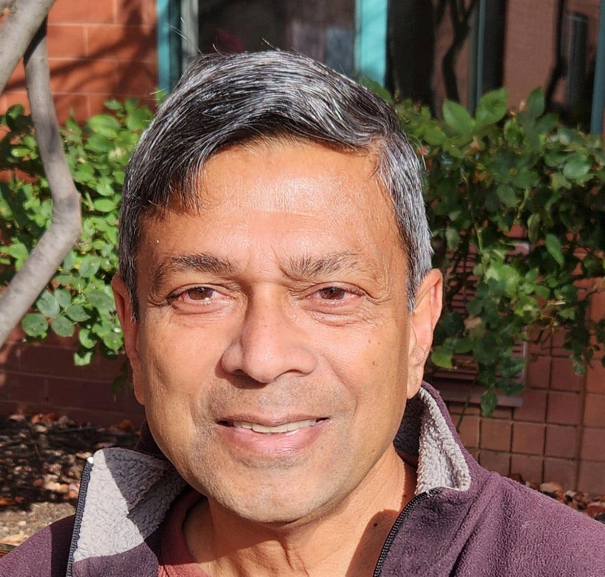 Headshot of Bangladeshi author Mahmud Rahman wearing a fleece jacket, smiling in front of sunlit foliage and a brick wall in the background.