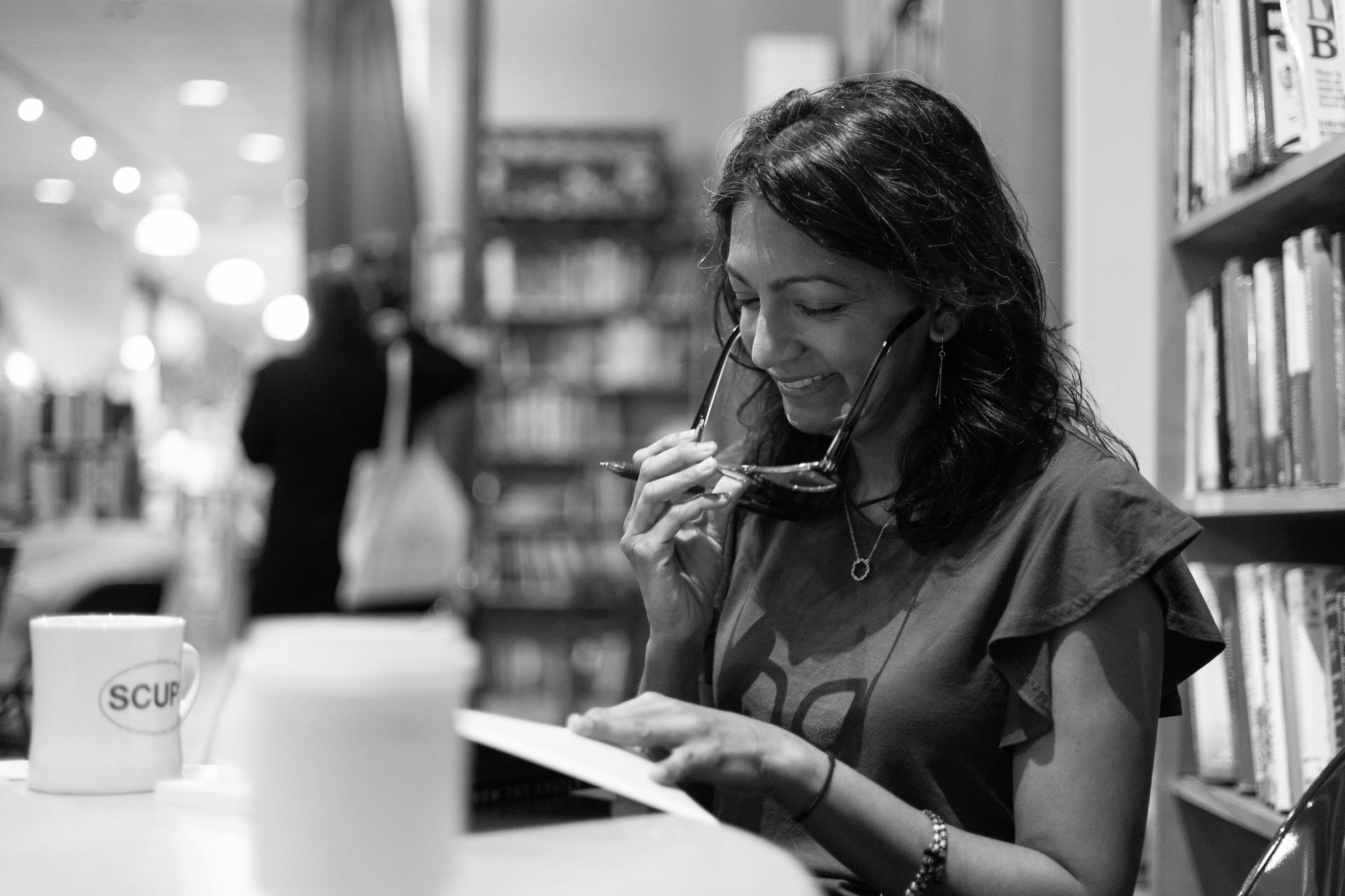 Bangladeshi American author Diruba Ahmed uses one hand to take off her glasses while holding a pen and the other hand to keep a book open on a table.