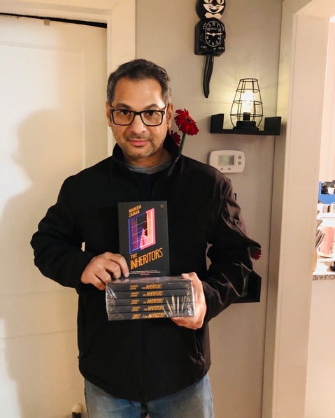 Bangladeshi American English professor and author Nadeem Zaman holds a stack of his books "The Inheritors" wearing glasses and a jacket inside a home.