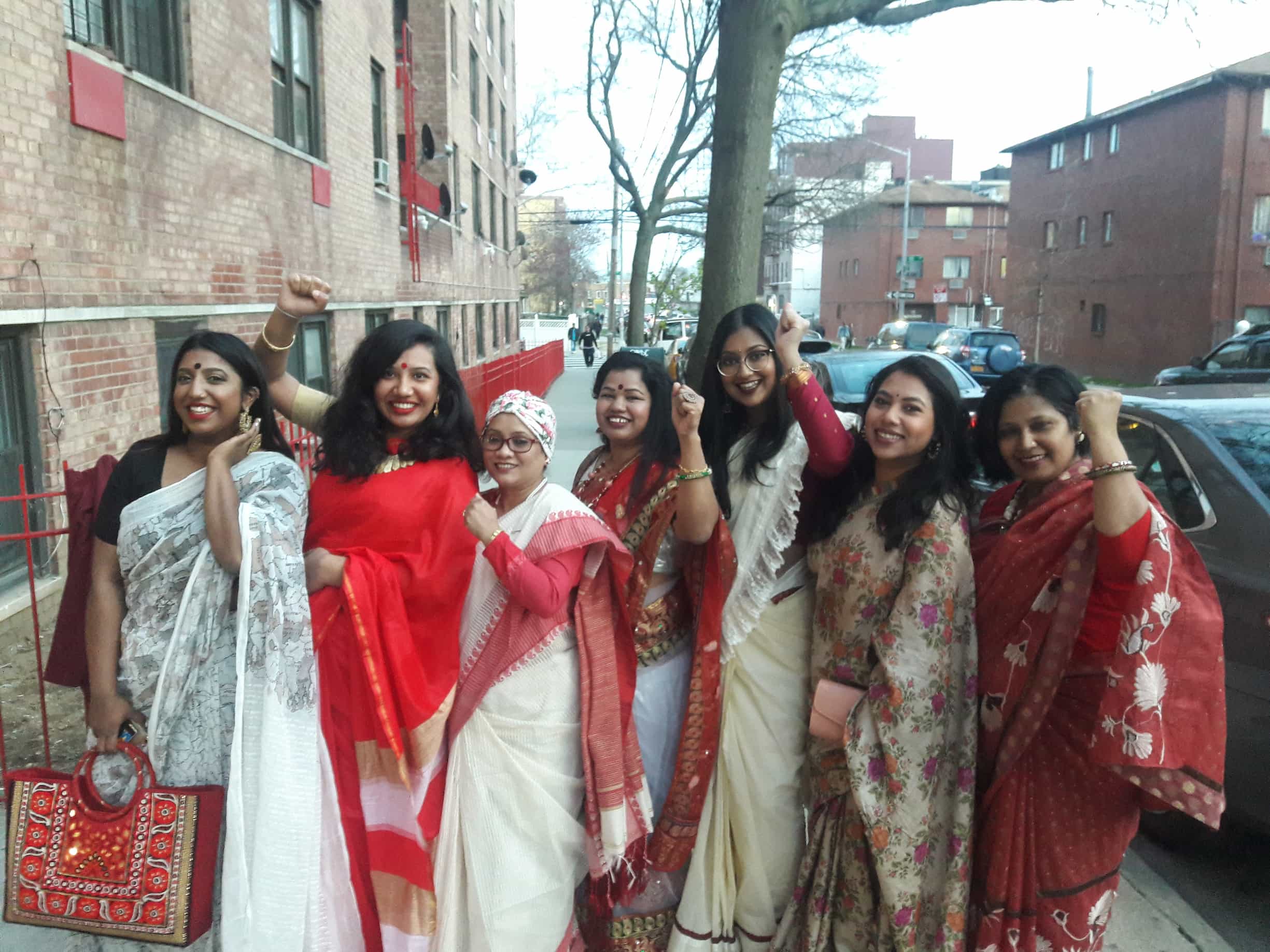 Seven Bangladeshi American female organizers pose wearing saree on a sidewalk in New York, surrounded by brick buildings and parked cars.