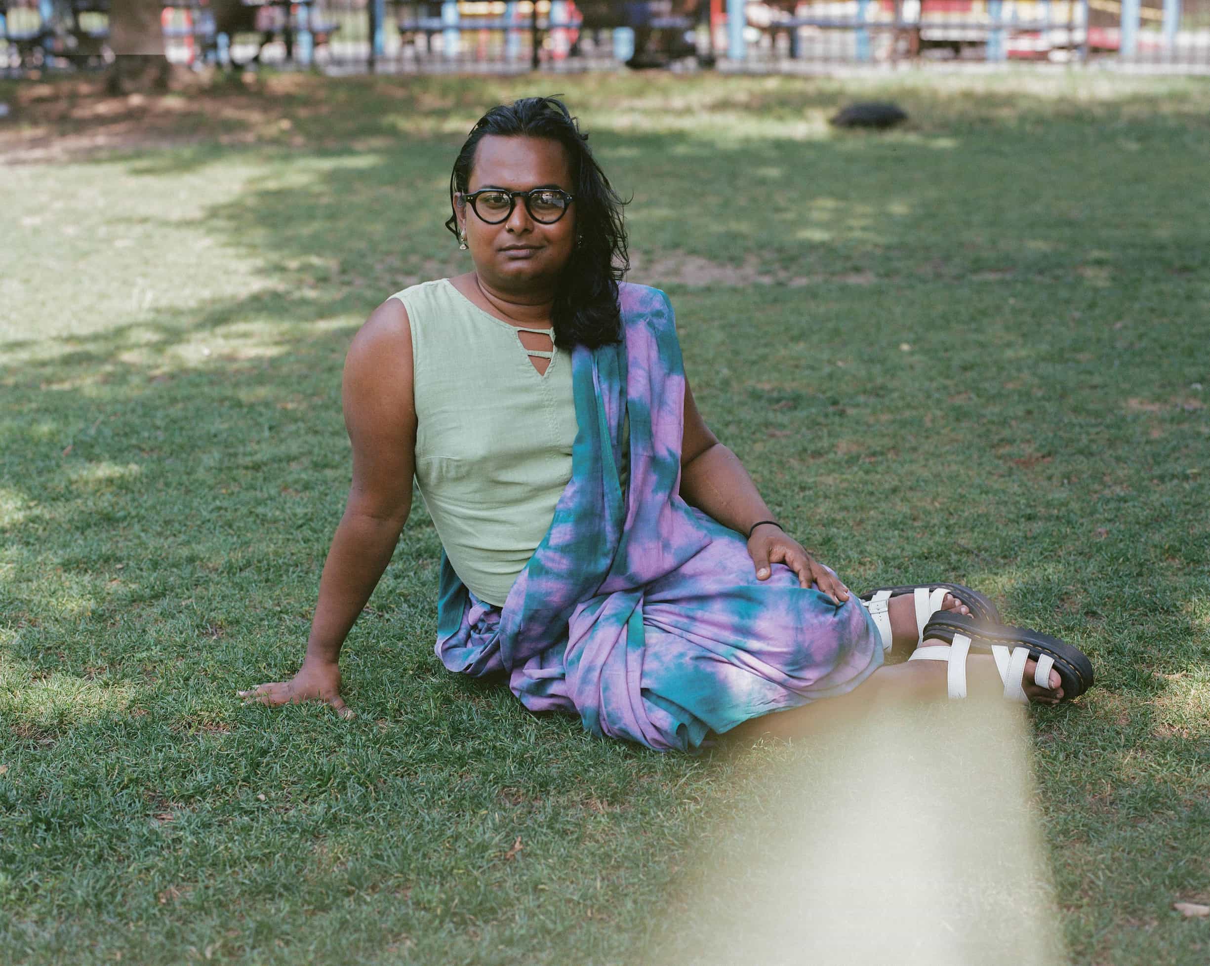 Bangladeshi artist Tara Asgar sits on the lawn with one arm propped to the side and the other arm resting on her leg while wearing a tie dye saree.