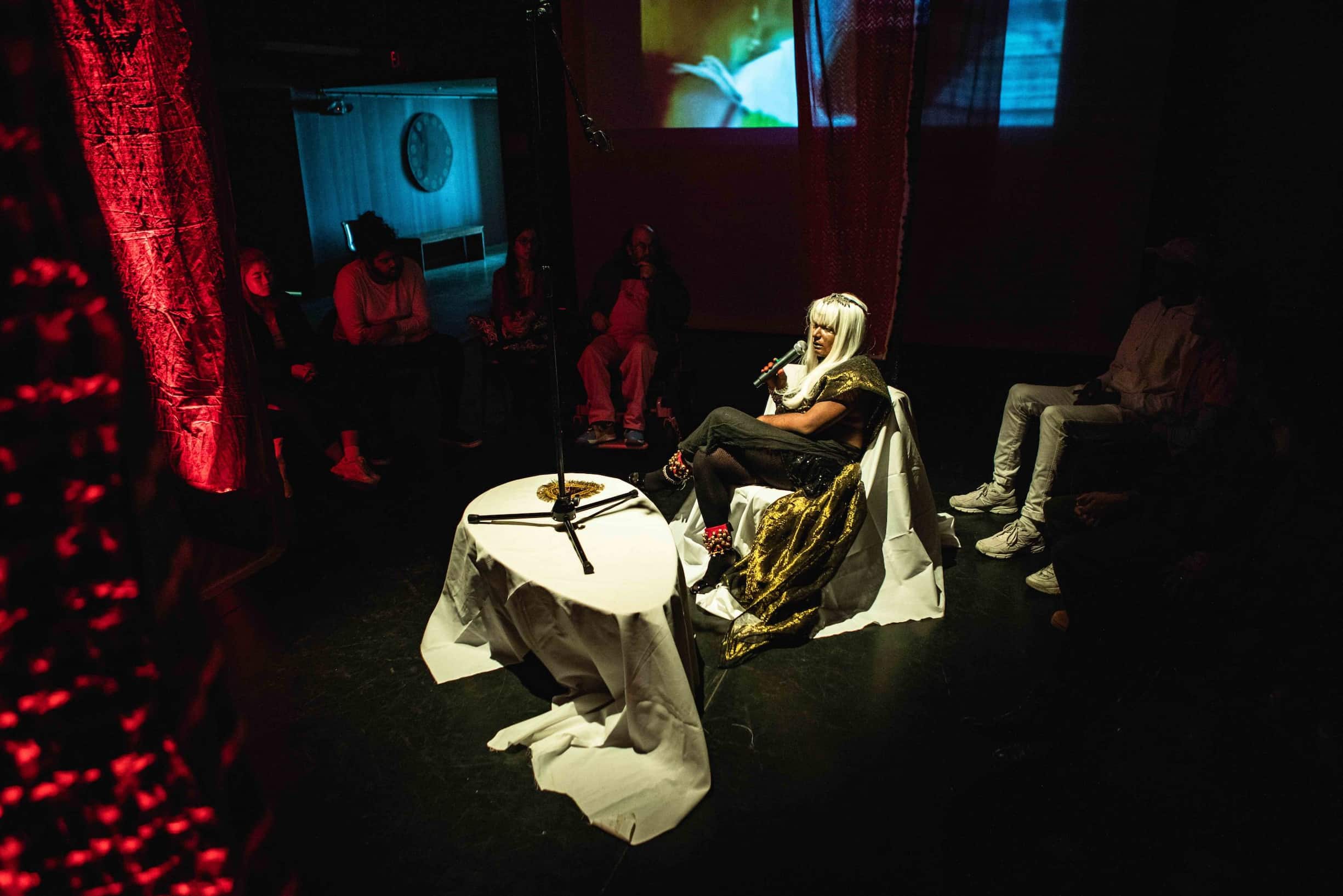 Bangladeshi artist Tara Asgar wearing a long wig is seated under the spotlight with a microphone in hand and surrounded by people sitting in a circle.