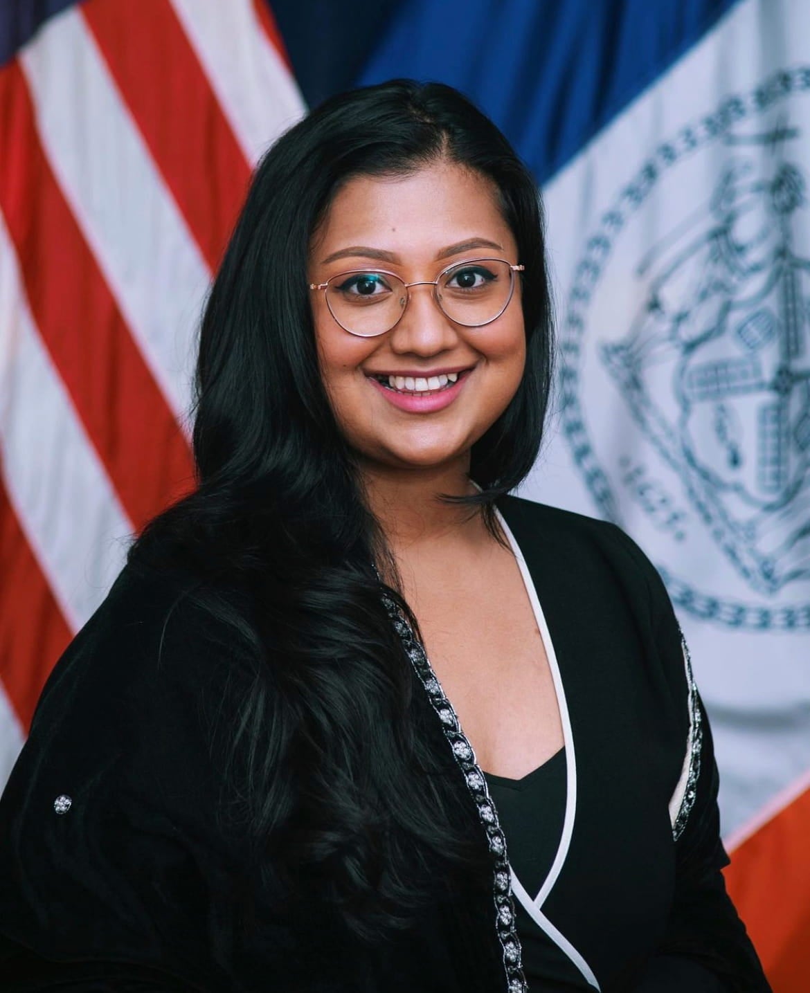 Headshot of Bangladeshi American Shahana Hanif wearing glasses and dressed in a wrap-style top, in front of an American flag and New York City flag.