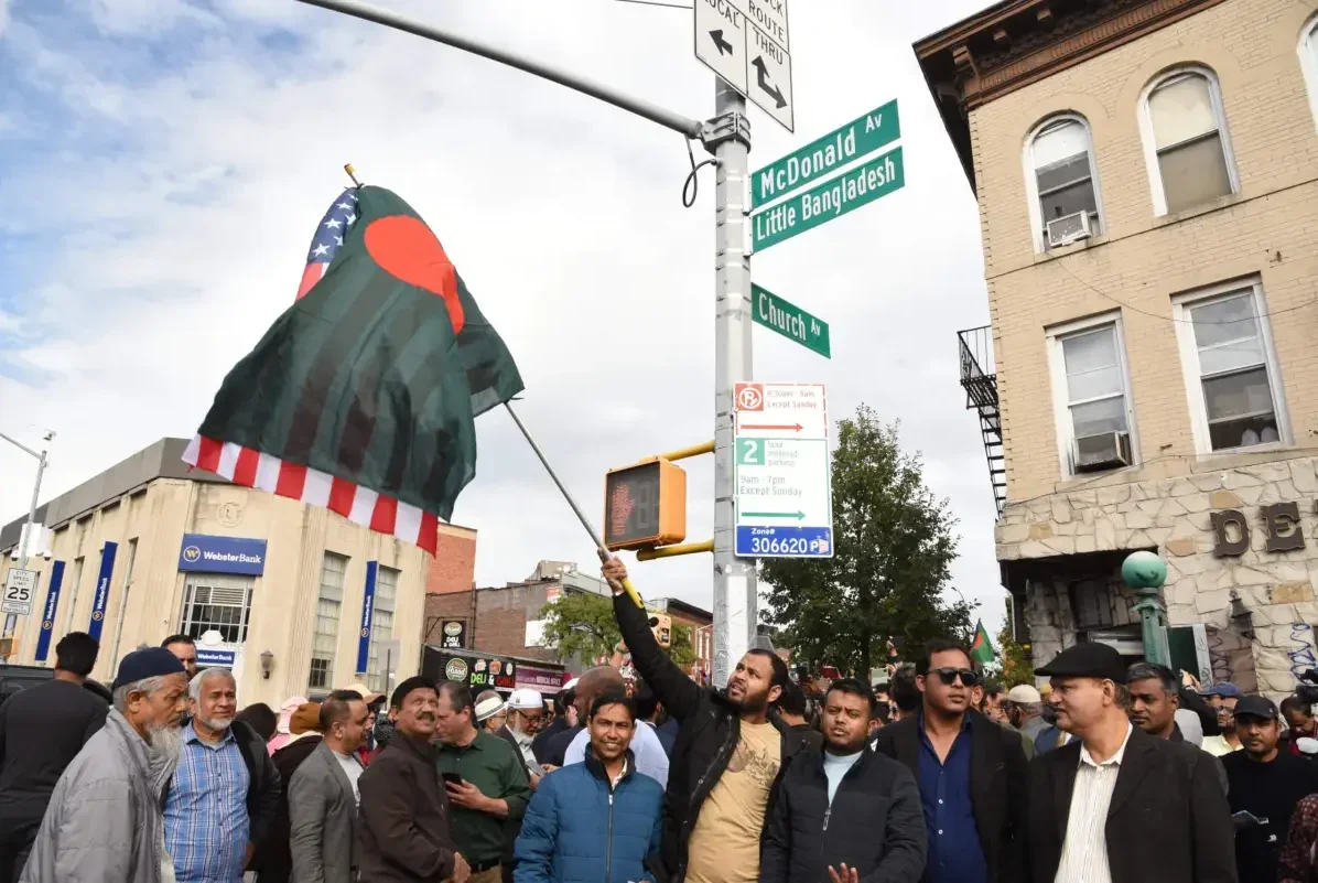 Bangladeshi Americans gather on the street corner to celebrate the naming of Little Bangladesh. A man holds a Bangladeshi flag and an American flag.