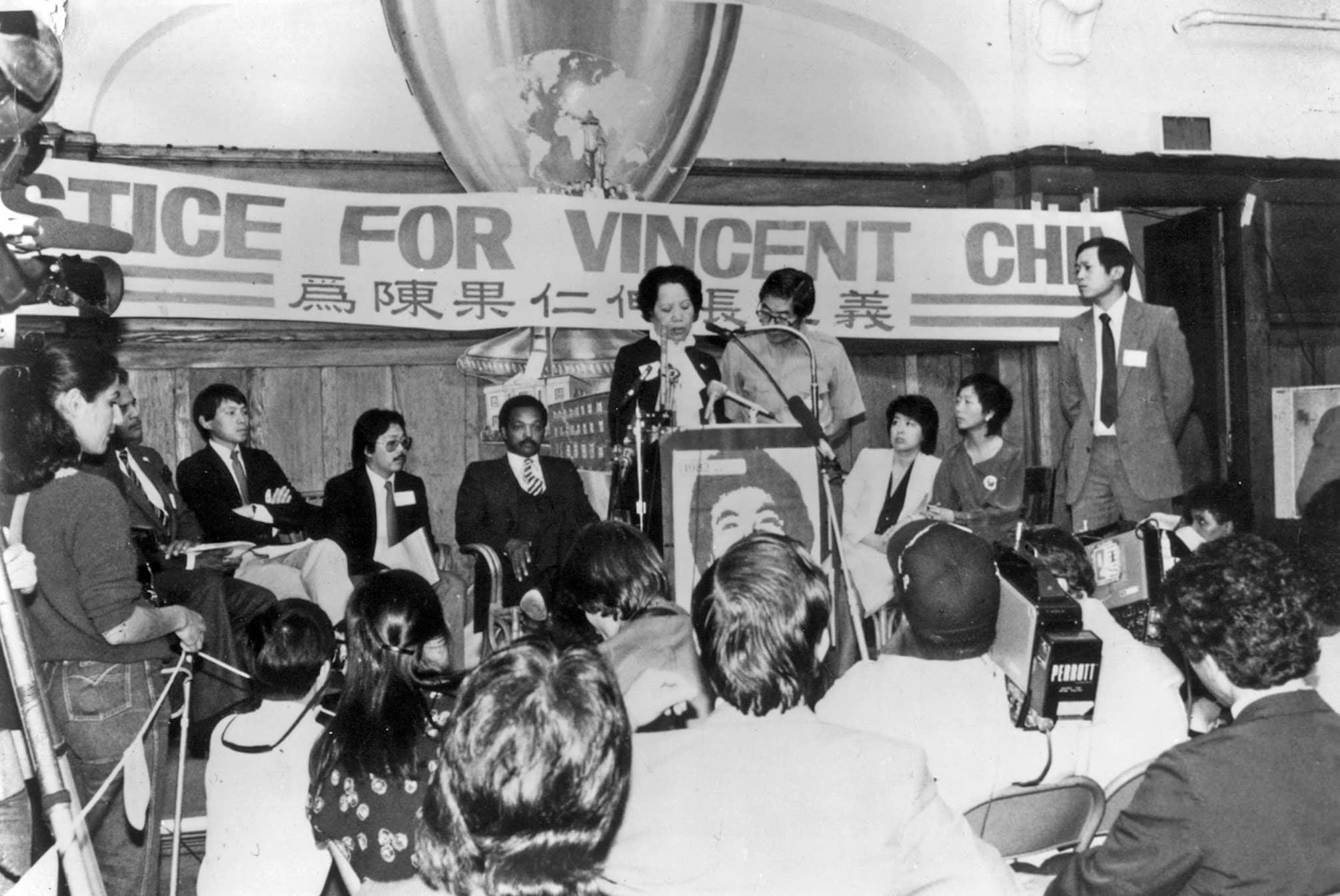 Lily Chin at podium in front of "Justice for Vincent Chin" banner and addresses audience. Seated behind her is Jesse Jackson, a Black man in suit.