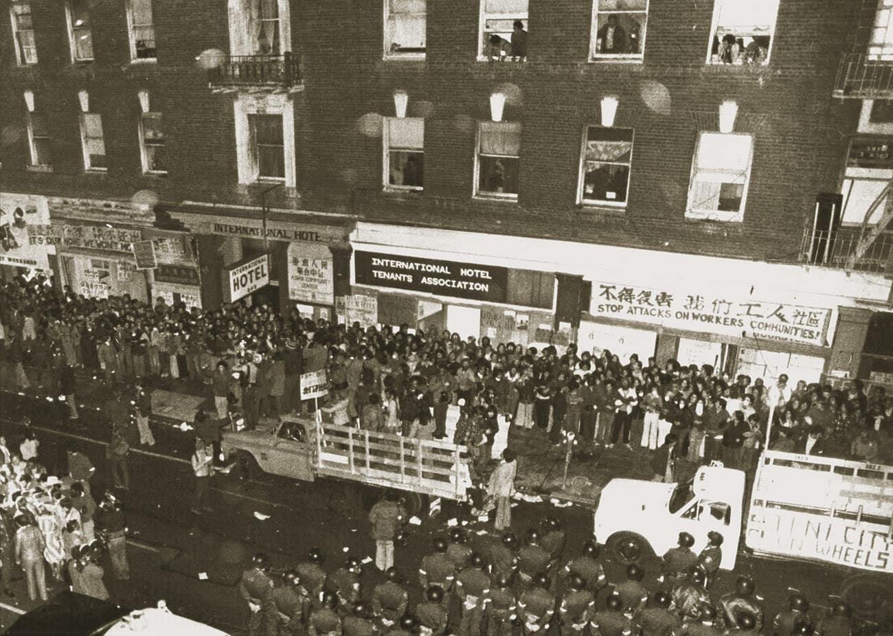 Aerial view of street in front of the I-Hotel. Rows of supporters form human barricade in front of the building. Rows of police stand opposite them.