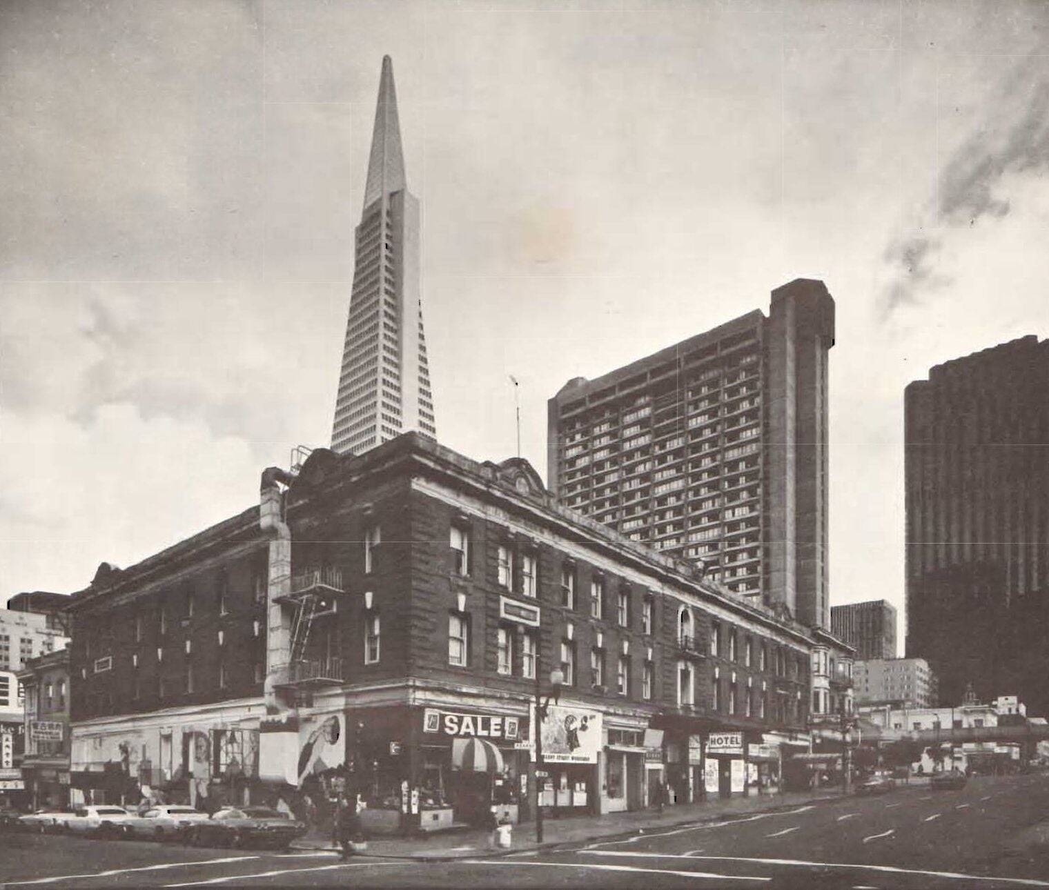 Exterior of the International Hotel, a low-rise brick building near intersection of two streets. Behind the hotel are several high-rise buildings.