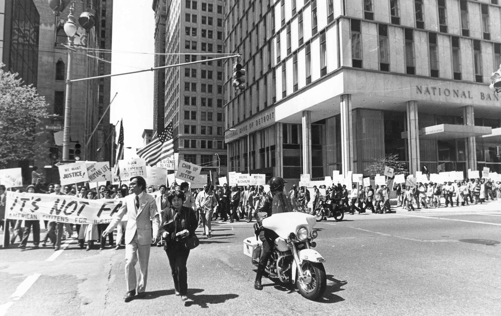 Large crowd marches with signs and banner. Crowd moves along sidewalk and through intersection. Police motorcycle rides beside two marchers at front.