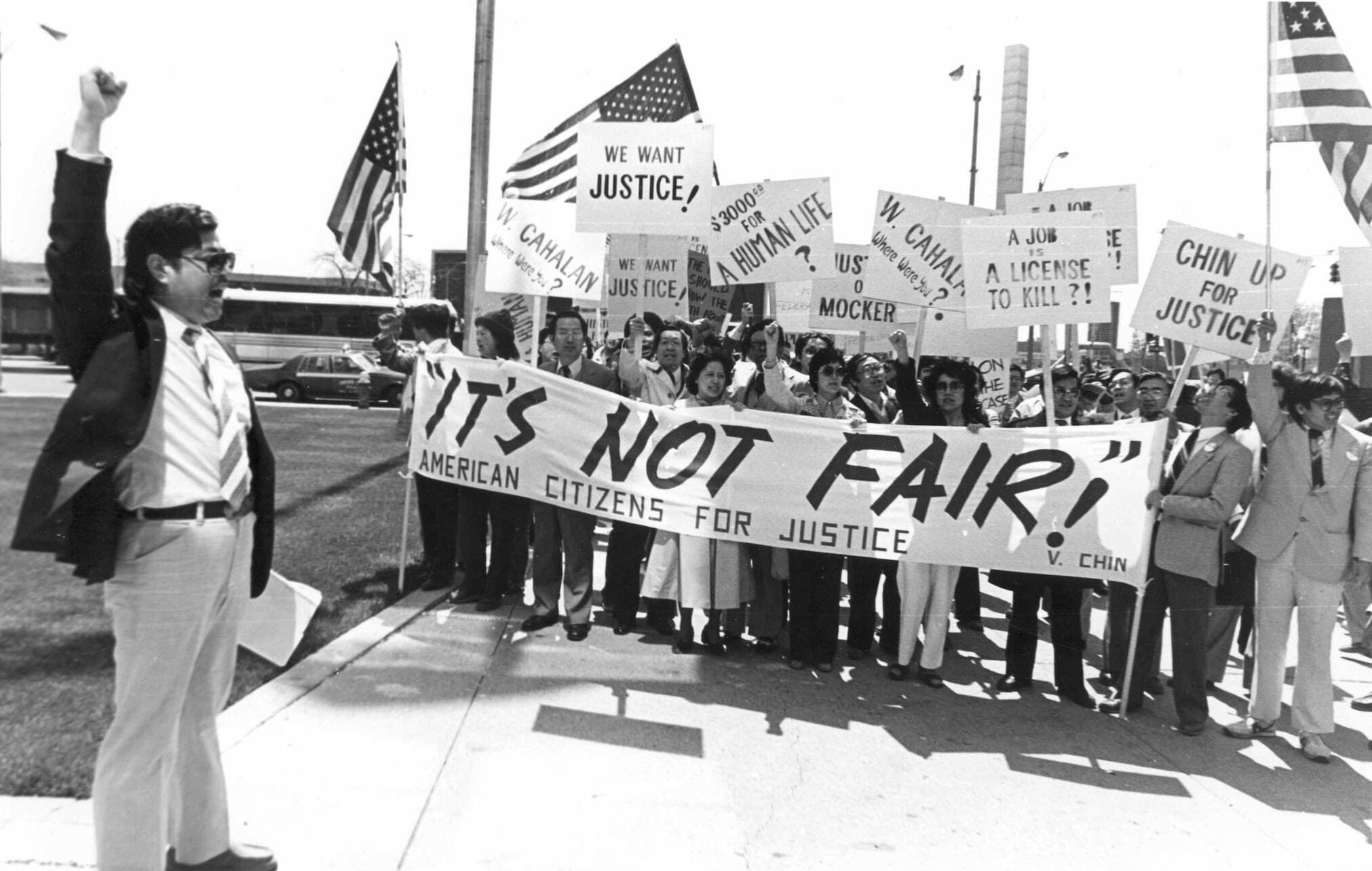 Man in suit jacket stands with raised fist before group of Asian Americans. They stand behind "It's Not Fair" banner and carry signs and U.S. flags.
