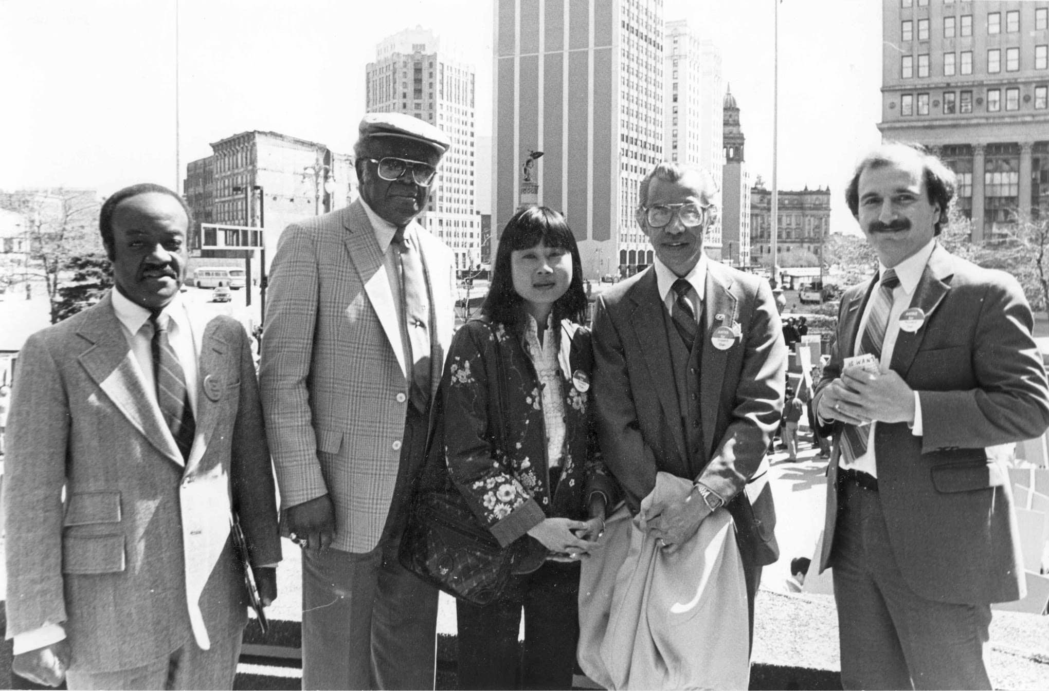Five people wearing pins reading "Justice for Vincent Chin" stand in a row for outdoor group photo with Detroit city skyline behind.