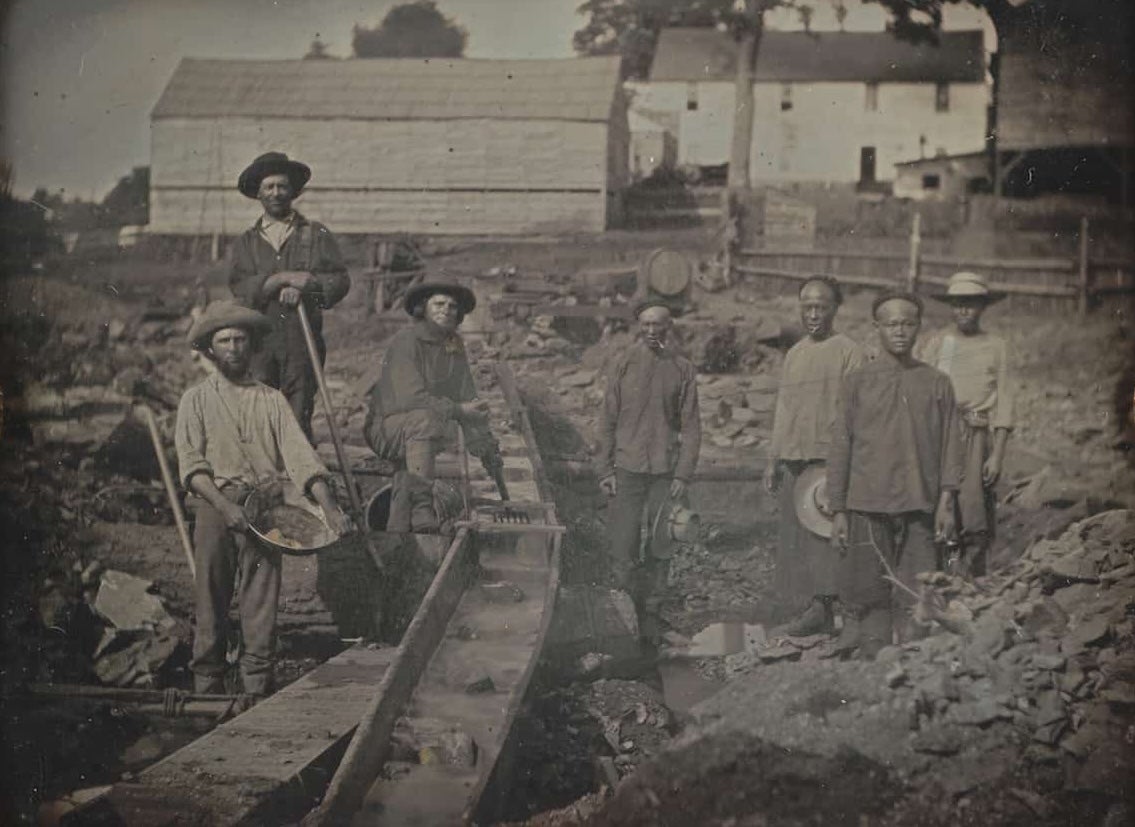 A photograph of a three white miners to the left of the sluice box and four Chinese miners to the right of the sluice box.