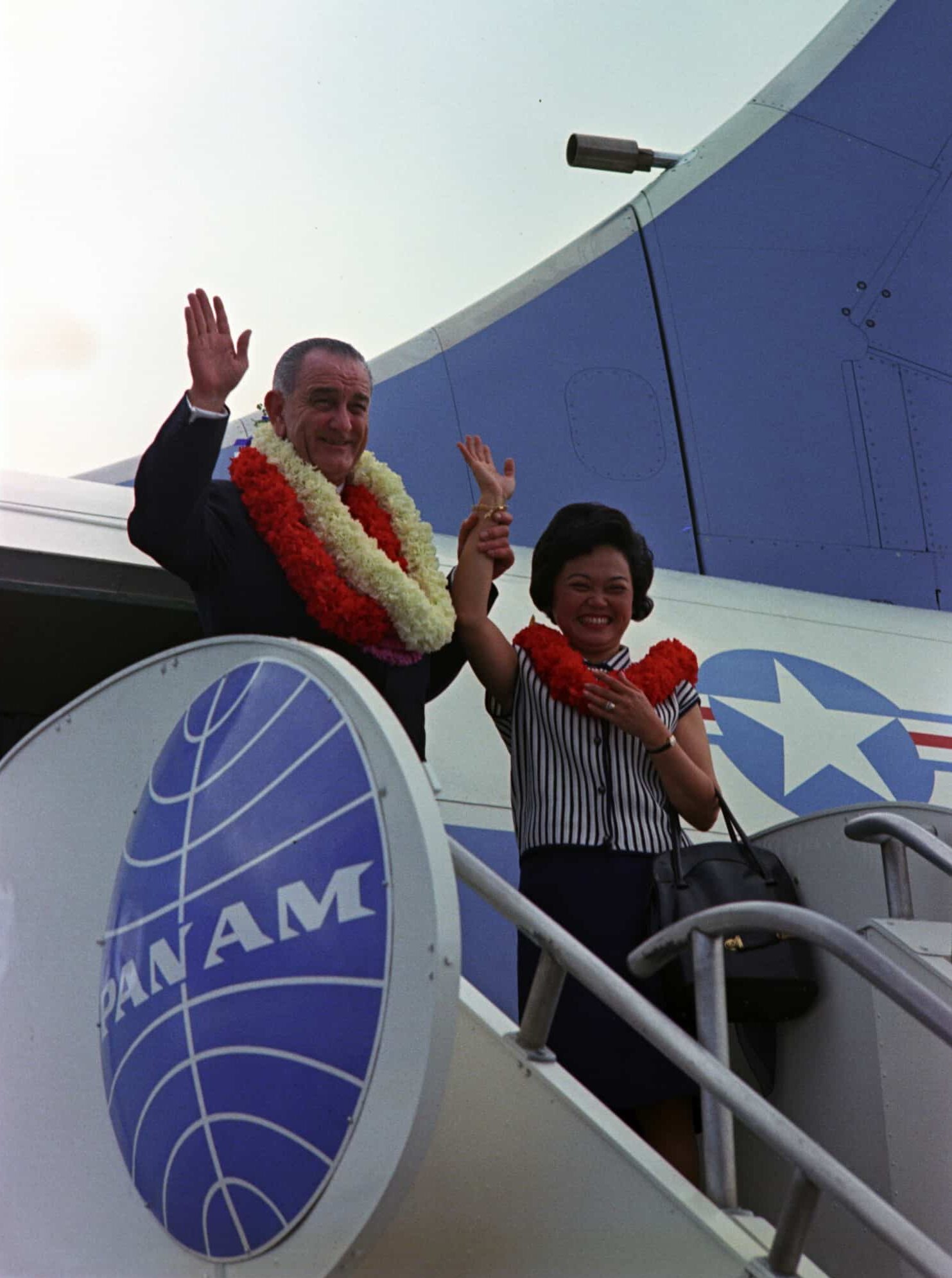 Patsy Mink, wearing striped shirt and red lei, waves to crowd from air stairs of plane. Next to her is President Johnson, in a suit with two lei.