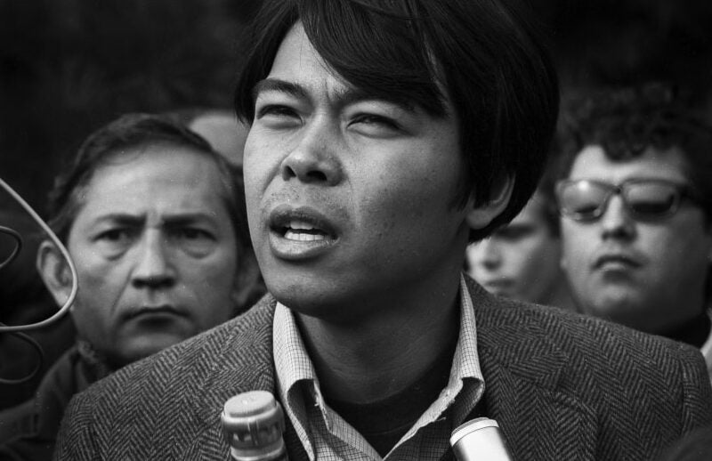 A close-up of Robert “Bob” Illumin, a young man in a patterned blazer and collared shirt, as he speaks into microphones. Crowd stands behind him.