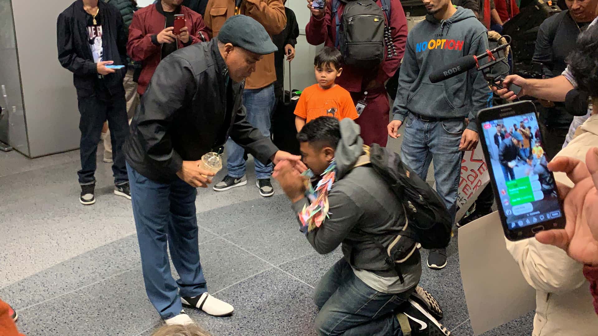 A crowd of cameras surround U.S. citizen Sok Loeun kneeling at his father's feet after returning to the U.S. from his self-deportation to Cambodia.