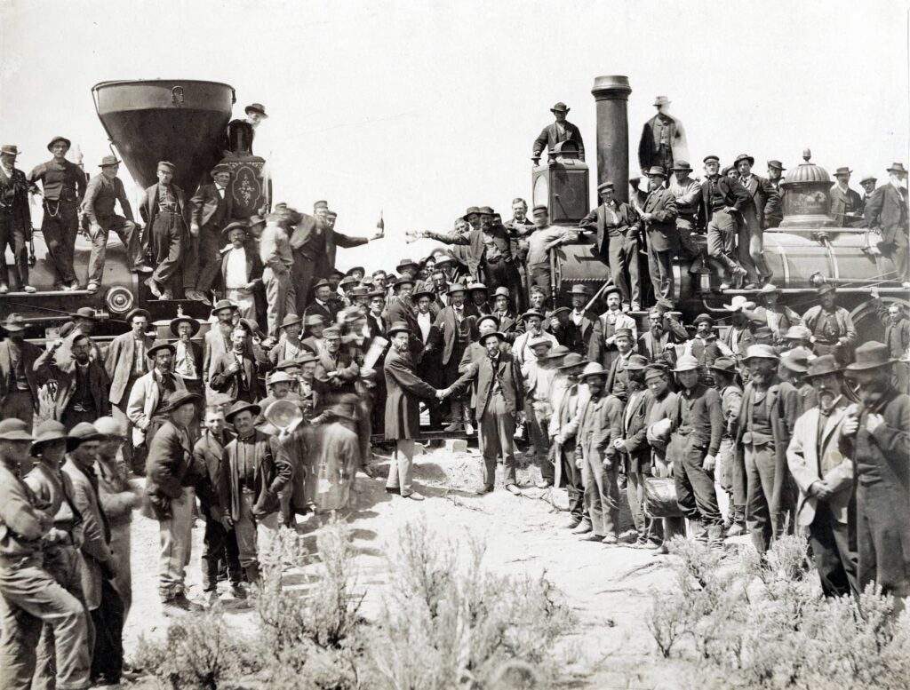 Two groups of railroad workers meet where the railroad tracks align. Two steam engines are nose to nose and engineers shake hands in the middle.