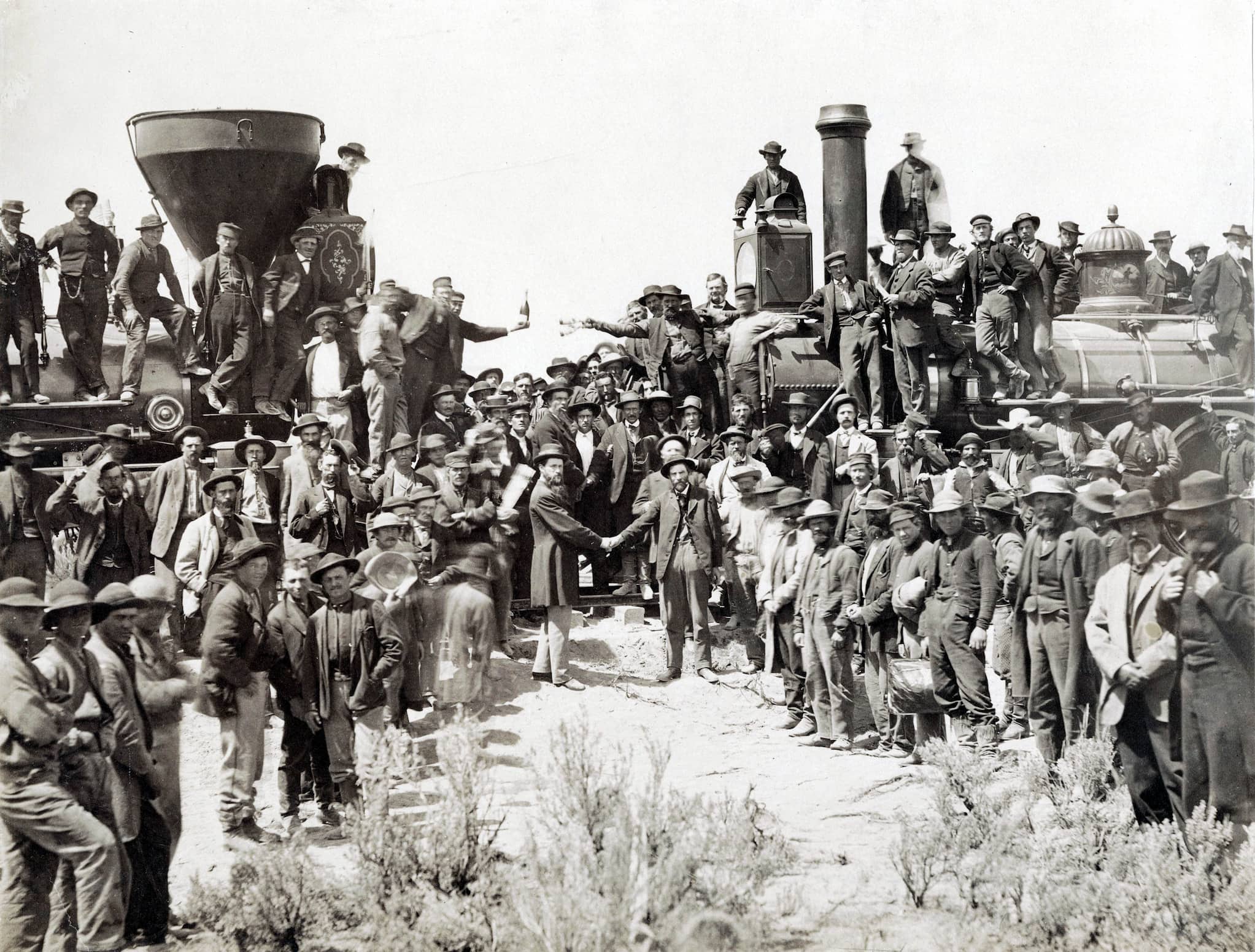 Two groups of railroad workers meet where the railroad tracks align. Two steam engines are nose to nose and engineers shake hands in the middle.